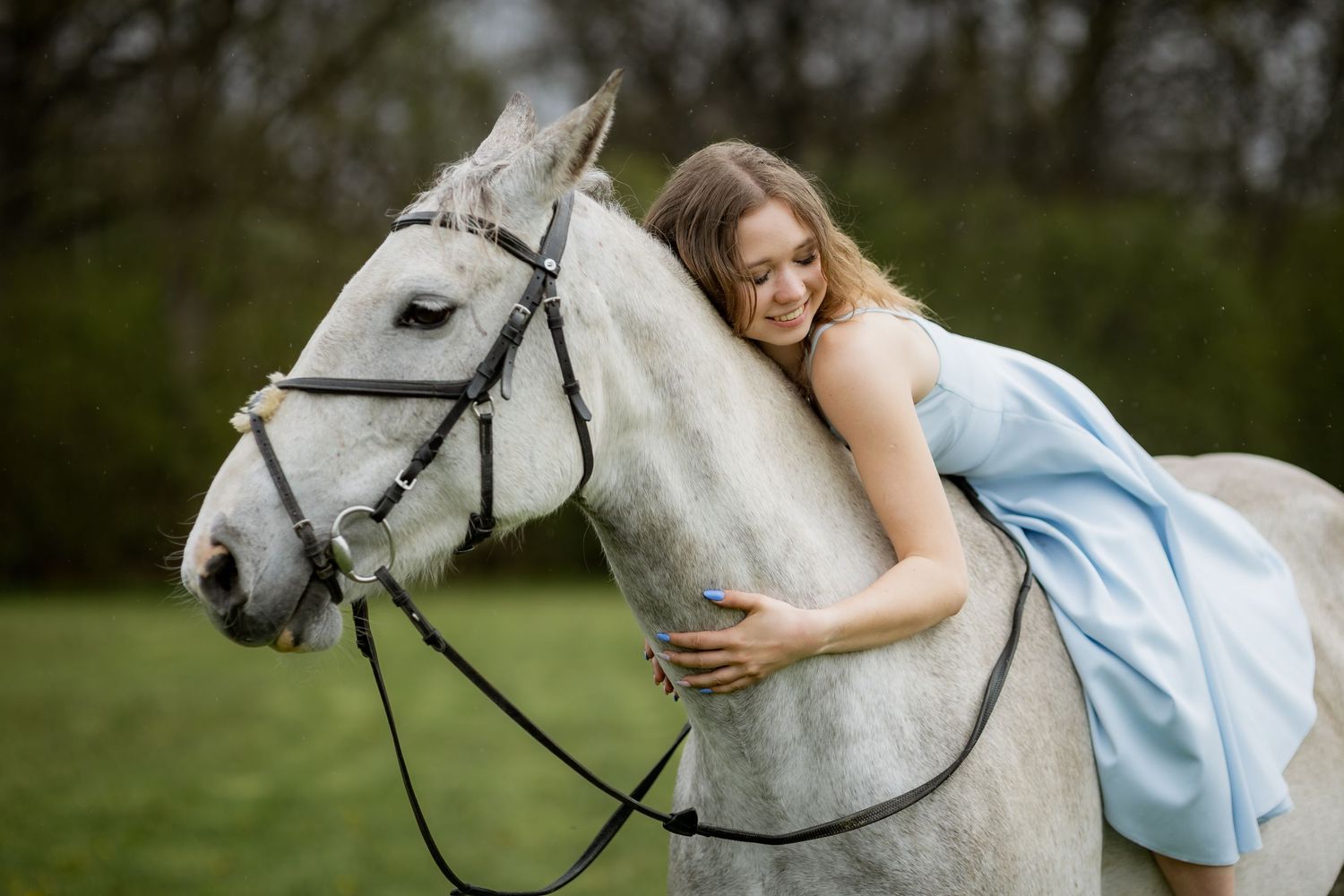 Young woman in blue dress leans forward hugging white horse affectionately in a park.