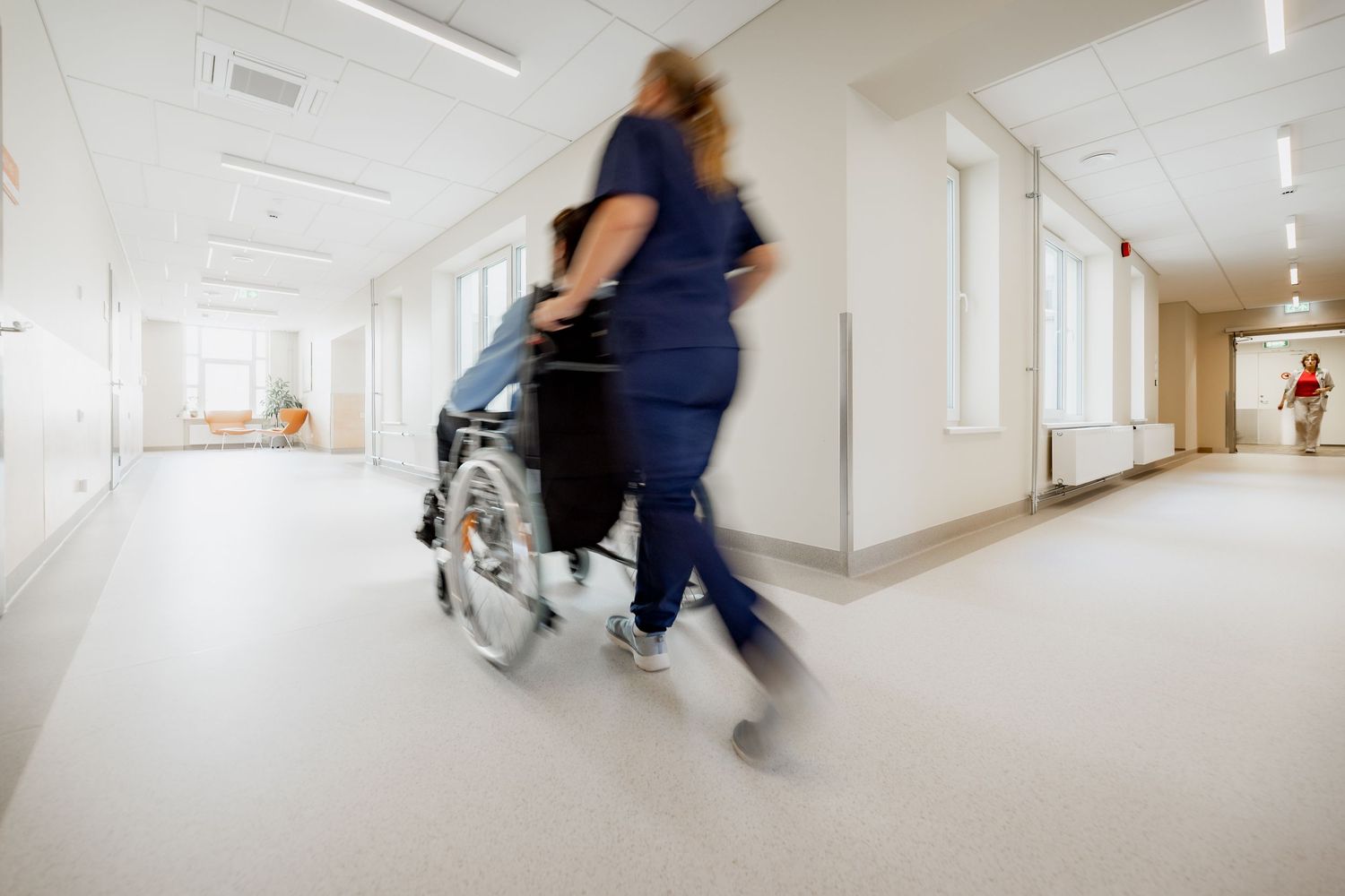 Nurse in blue scrubs urgently pushing patient in wheelchair down bright hospital corridor.