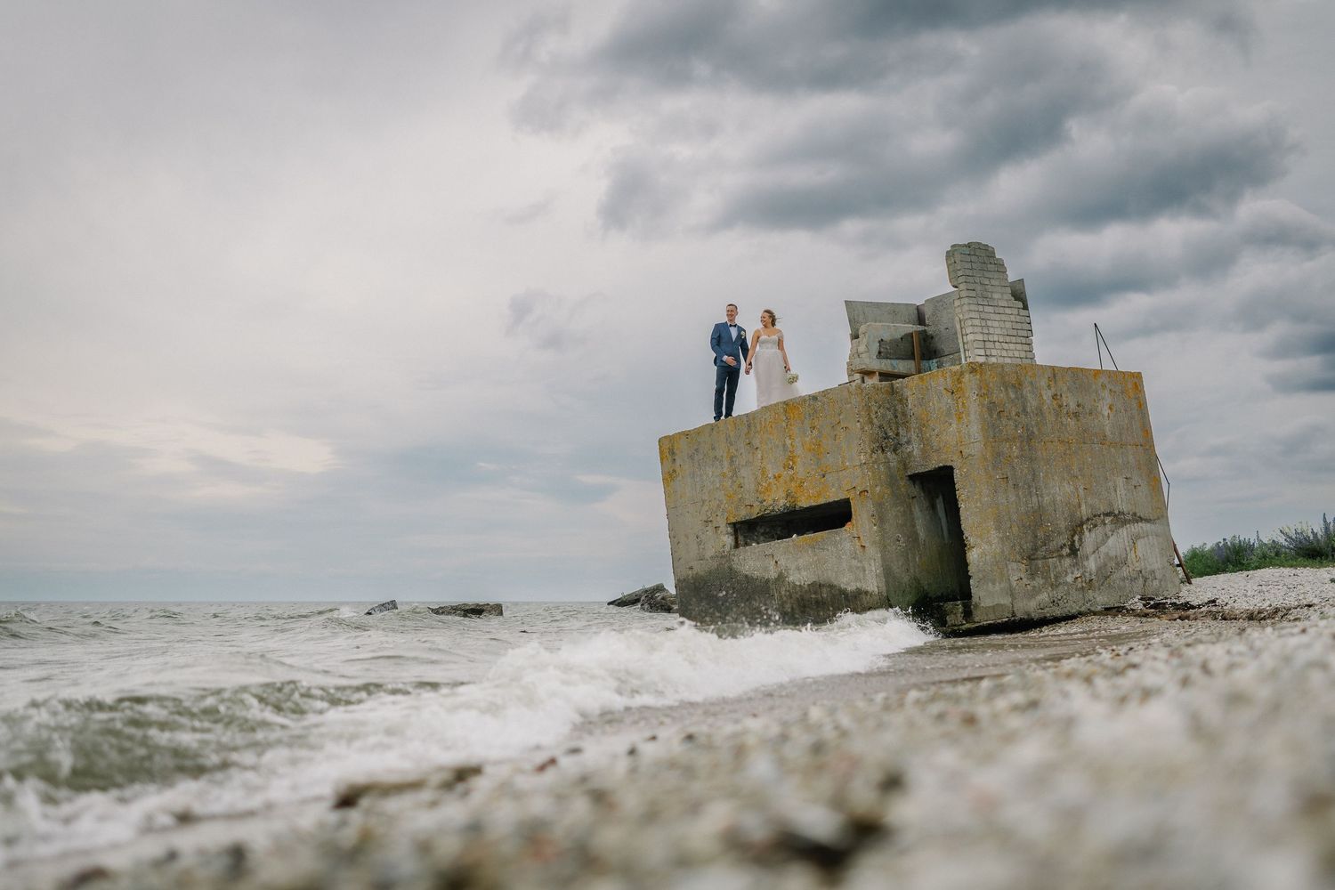 Couple standing atop a concrete bunker on a rocky beach with dramatic cloudy skies and ocean waves.