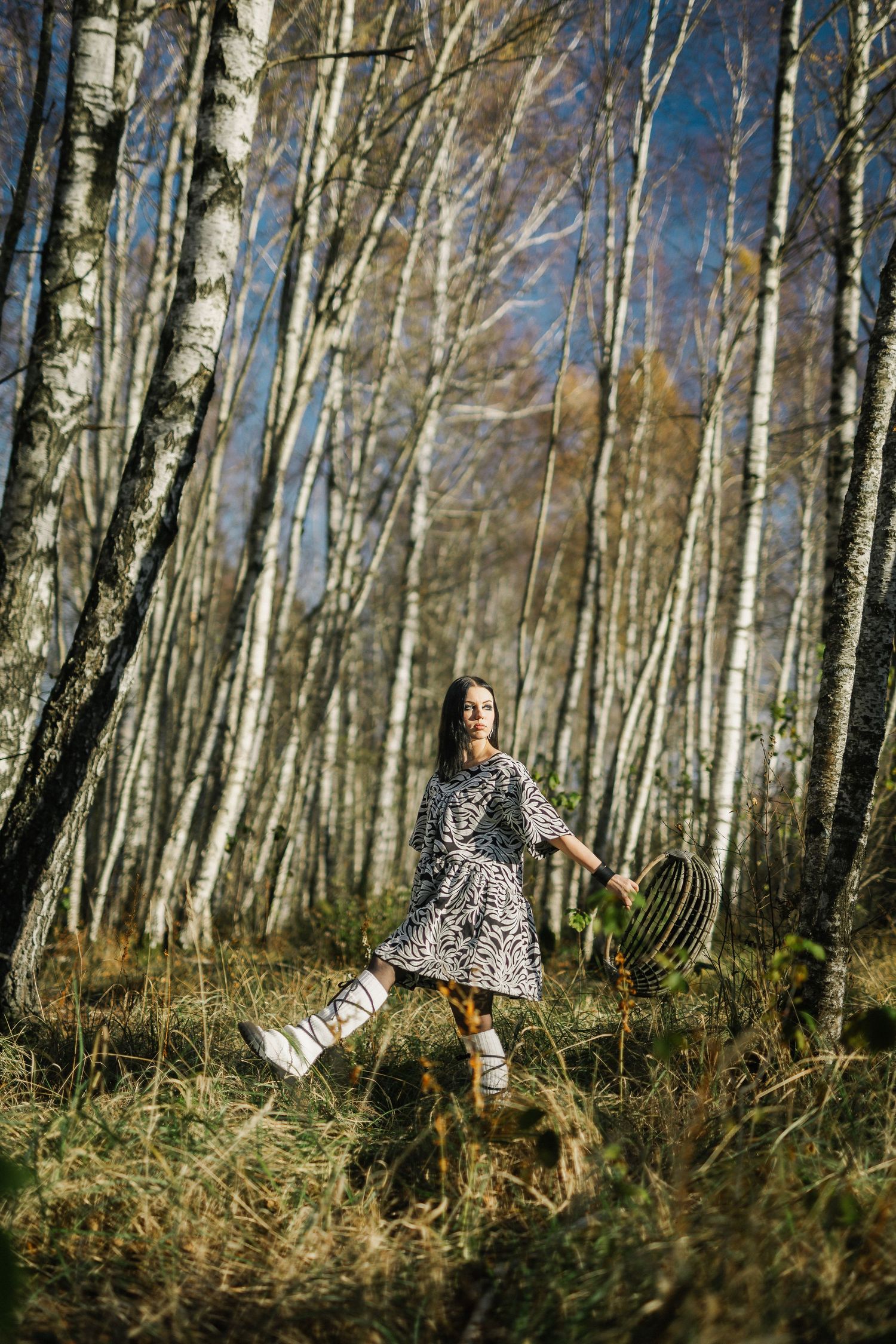Young woman in animal print dress holding a wicker basket walks through a sunny birch forest in autumn.