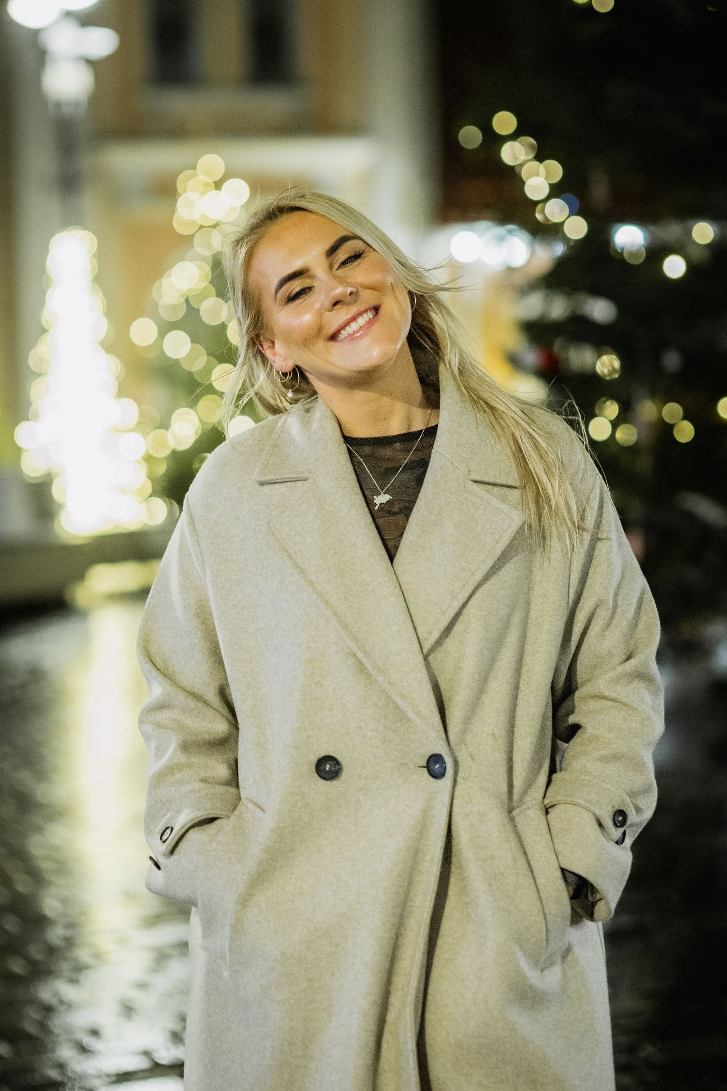 Happy blonde woman in beige coat smiles outdoors near glowing Christmas tree lights at night.