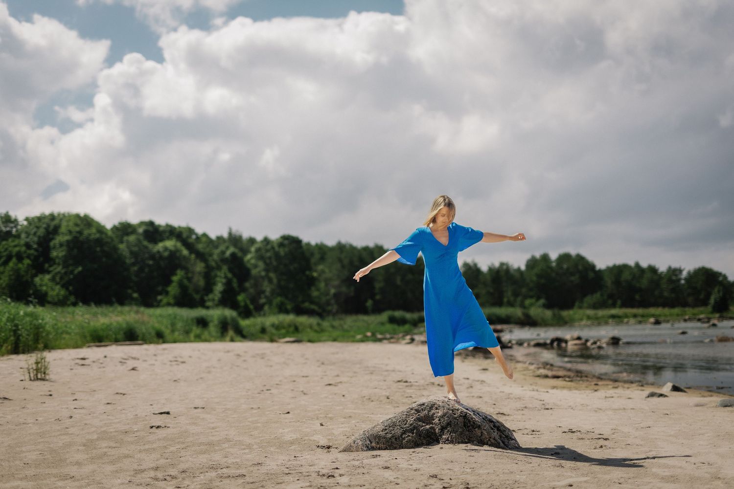 Woman in blue dress balances on rock at sandy riverbank with arms outstretched under cloudy sky.