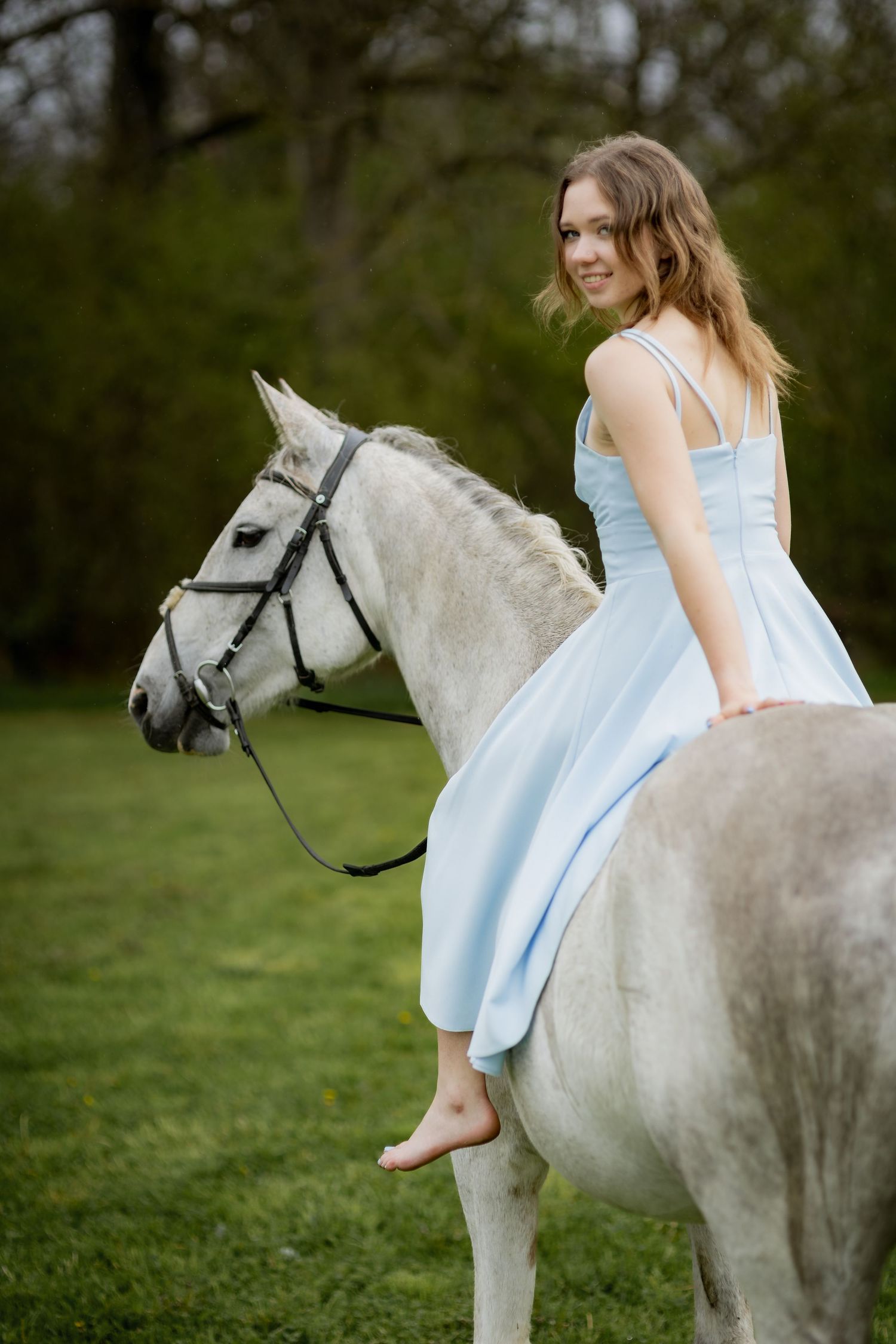 Smiling young woman in light blue dress rides white horse bareback in green outdoor setting.