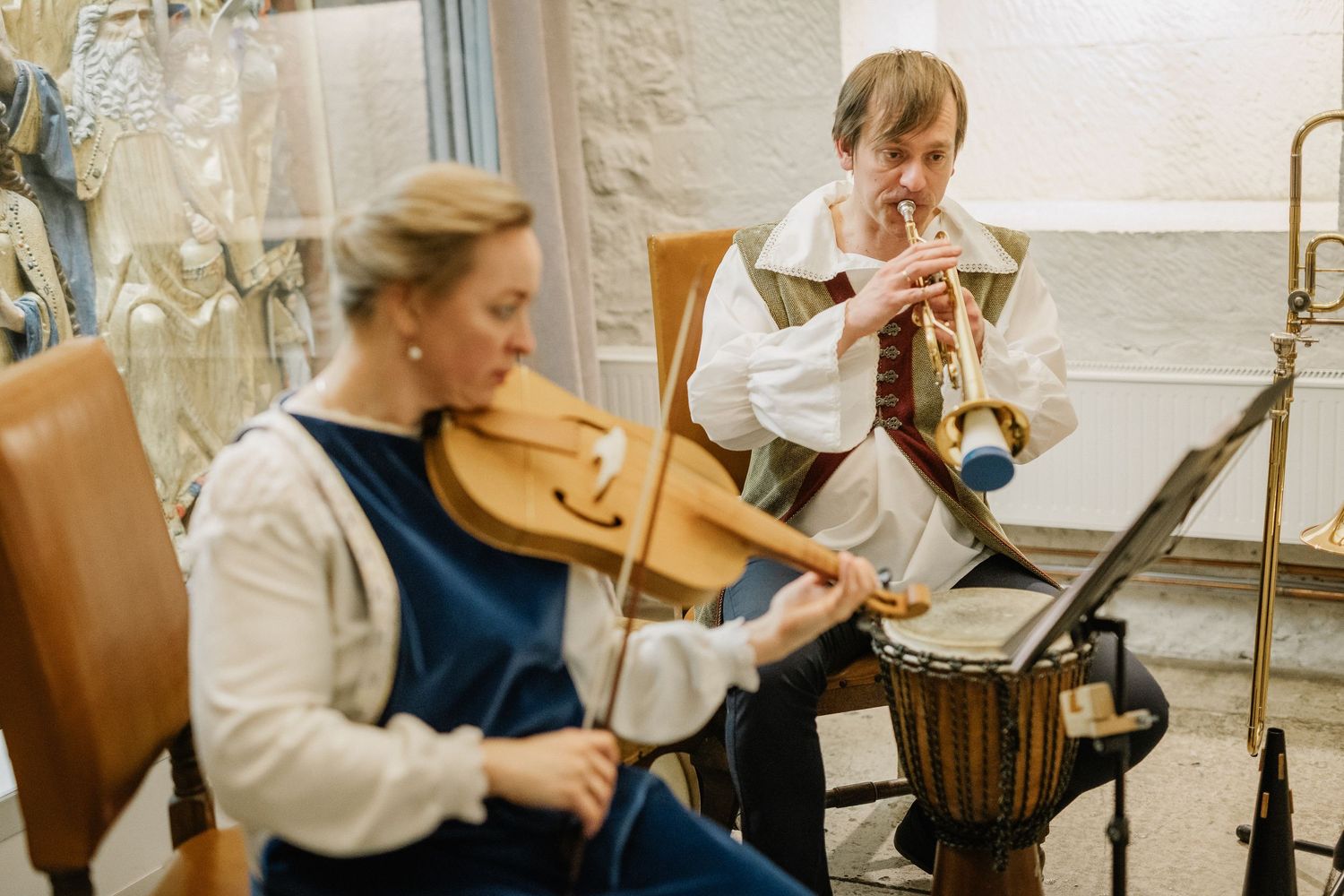 Two musicians in historical costumes play violin and trumpet in a rustic studio setting.