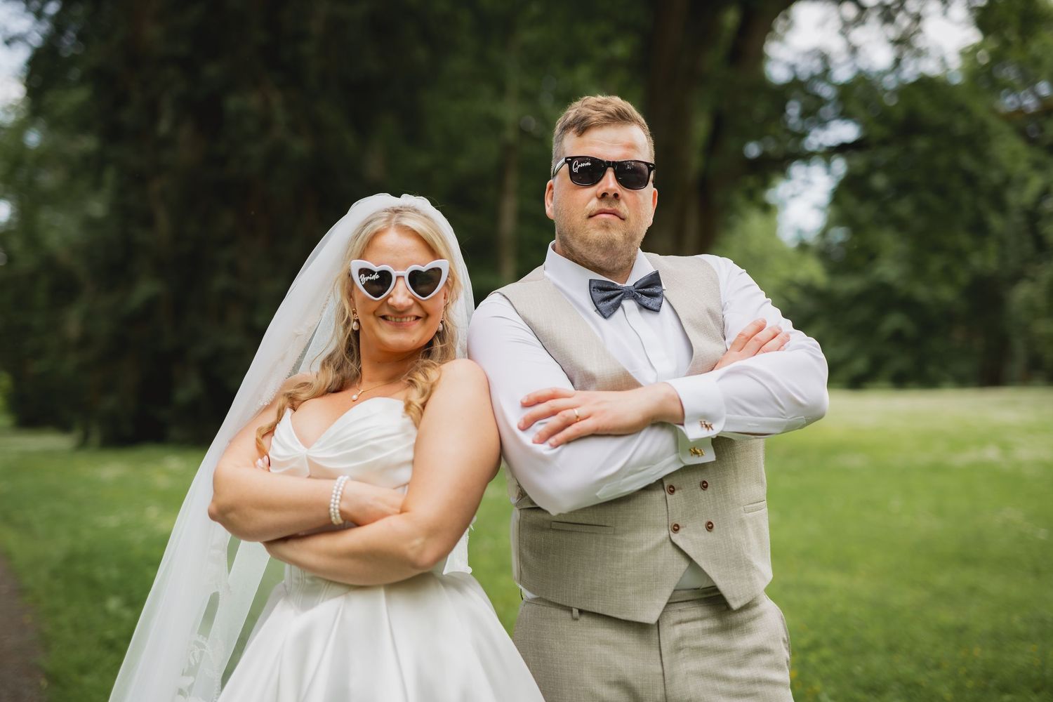 Bride and groom posing back-to-back with arms crossed wearing stylish sunglasses on their wedding day outdoors.