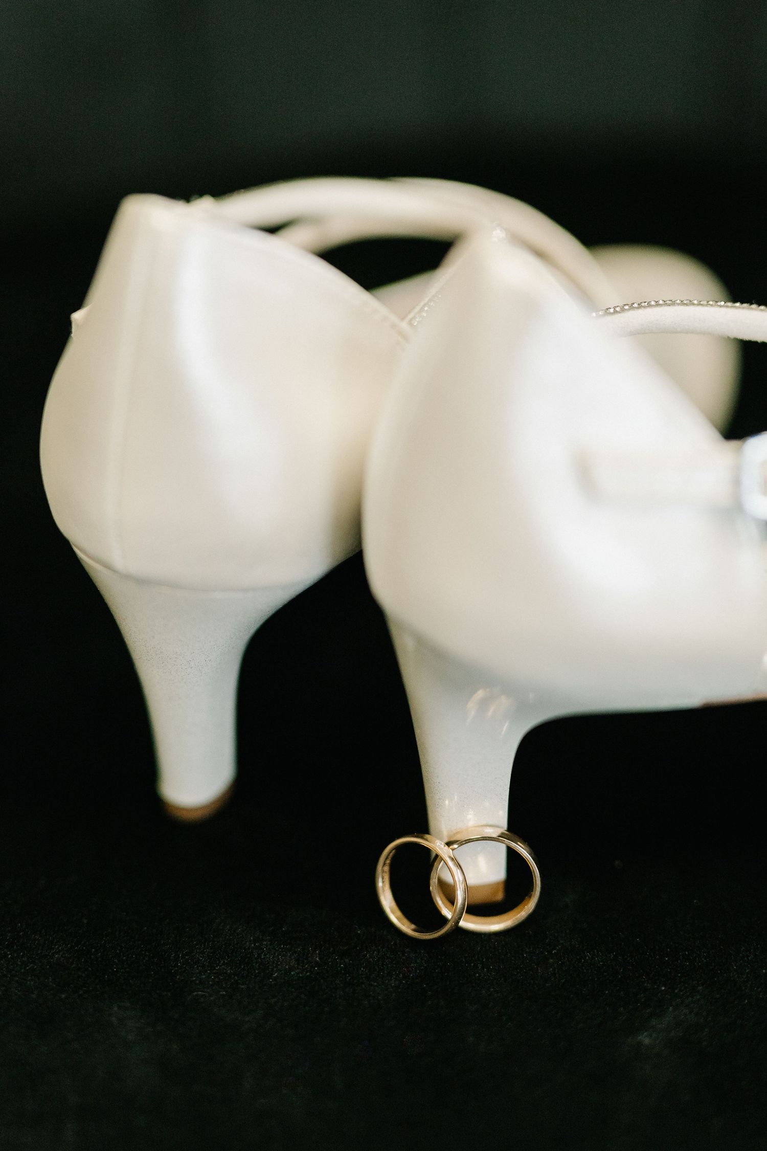 Two gold wedding rings rest between white satin bridal heels on a dark velvet surface.