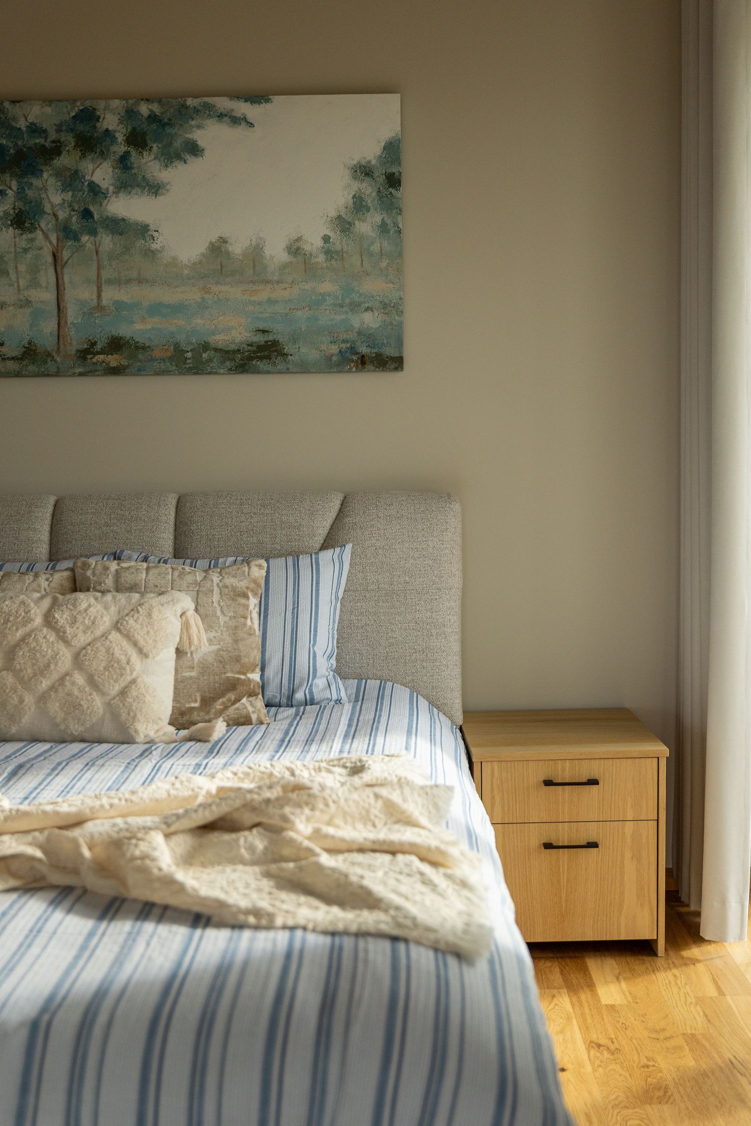 Cozy bedroom with blue striped bedding, textured pillows, wooden nightstand, and landscape painting above headboard.
