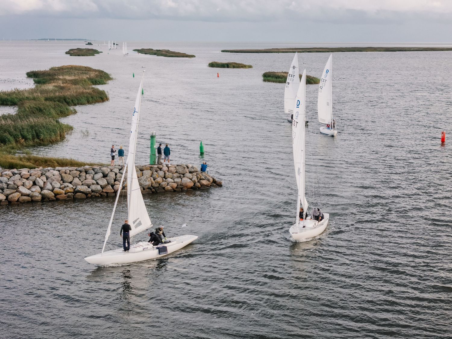 Two sailboats navigating calm coastal waters near a rocky shoreline with green flags and spectators watching nearby.