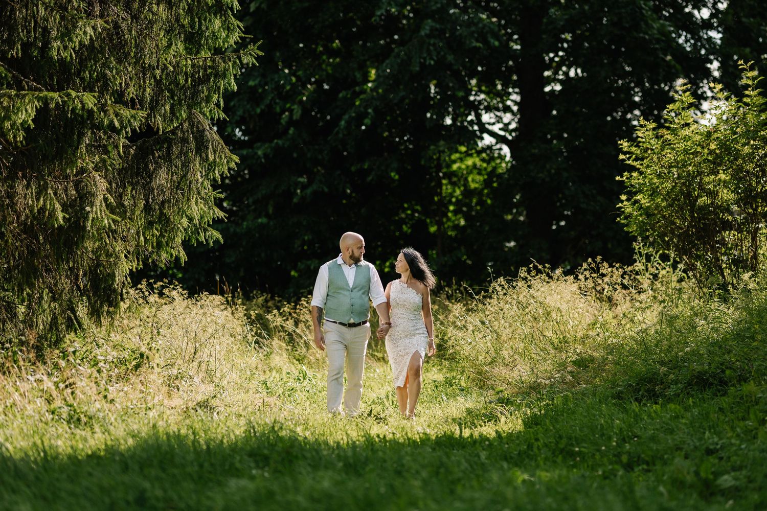 Couple holding hands walking through lush green meadow with trees, wearing elegant wedding attire on sunny day.
