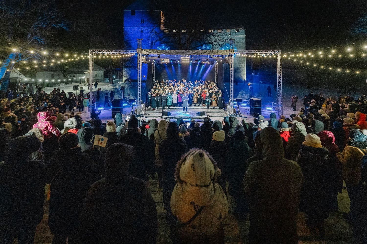 Outdoor winter concert at night with a lit stage, festive string lights, castle ruins backdrop, and a large crowd of spectators.