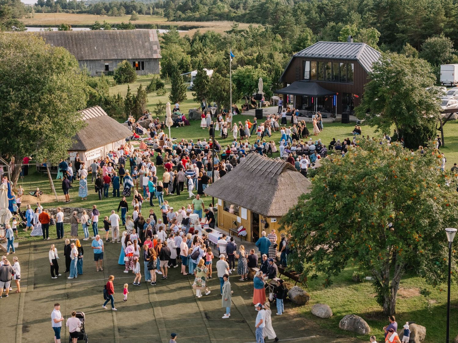 Aerial view of a large crowd gathered at a rural Estonian farm festival surrounded by trees and traditional buildings.