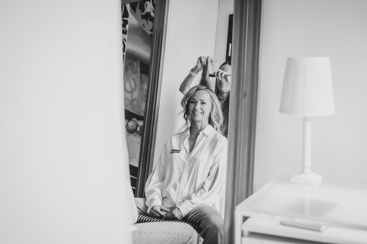 Black and white photo of a woman styling her hair while seated in front of a large mirror in a bright room.