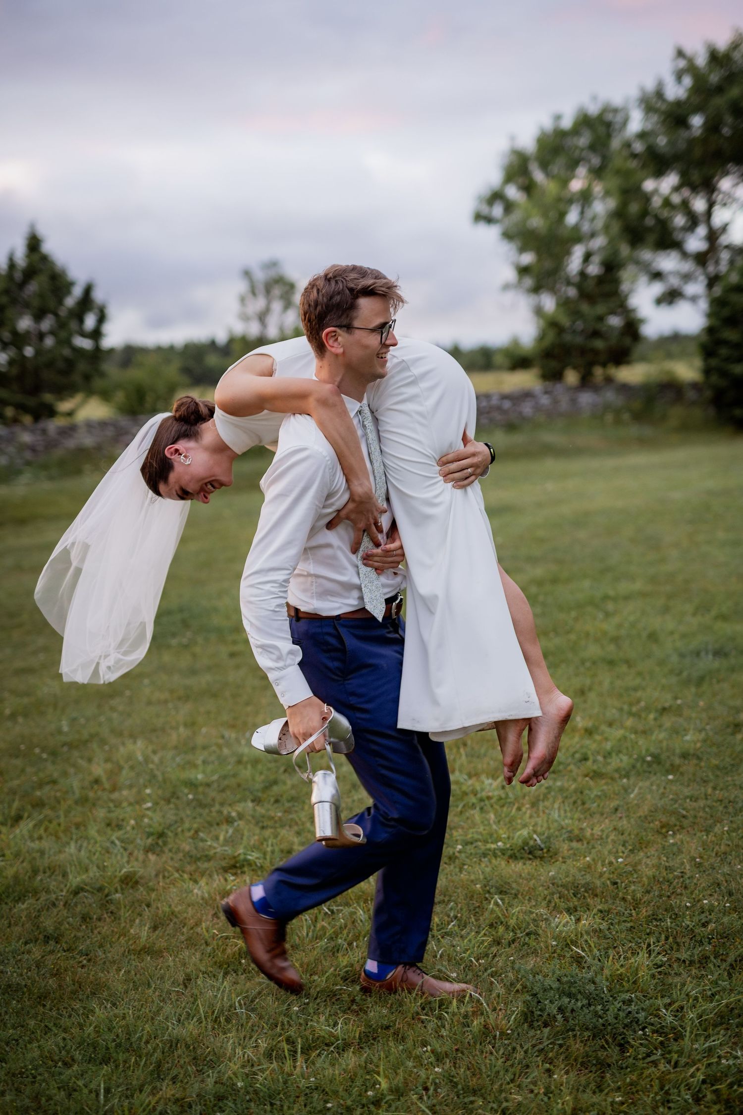 Groom carries laughing bride over his shoulder on green lawn at outdoor wedding reception at dusk.