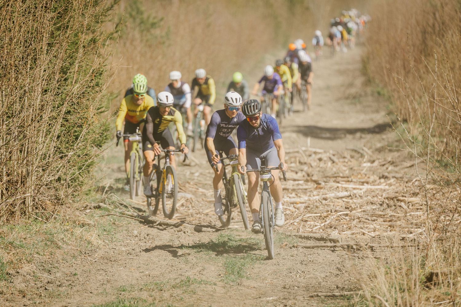 Cyclists racing on a dusty gravel road during a competitive cycling event, with a peloton of riders in colorful jerseys.