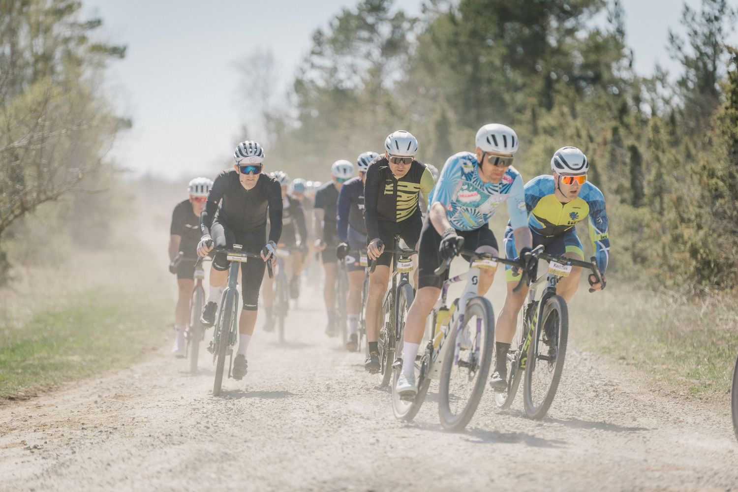 Cyclists race on a dusty gravel road during a competitive cycling event surrounded by trees.