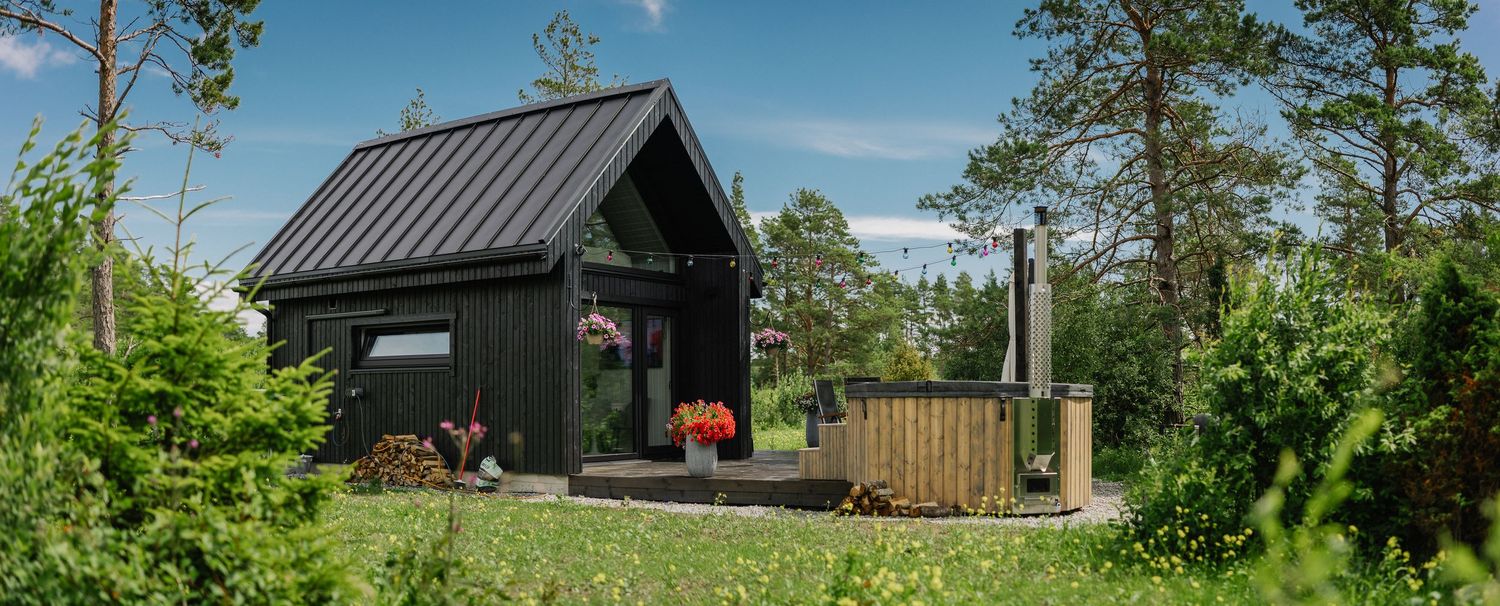 Black wooden cabin with metal roof surrounded by lush greenery, featuring a wooden hot tub and deck on a sunny day.