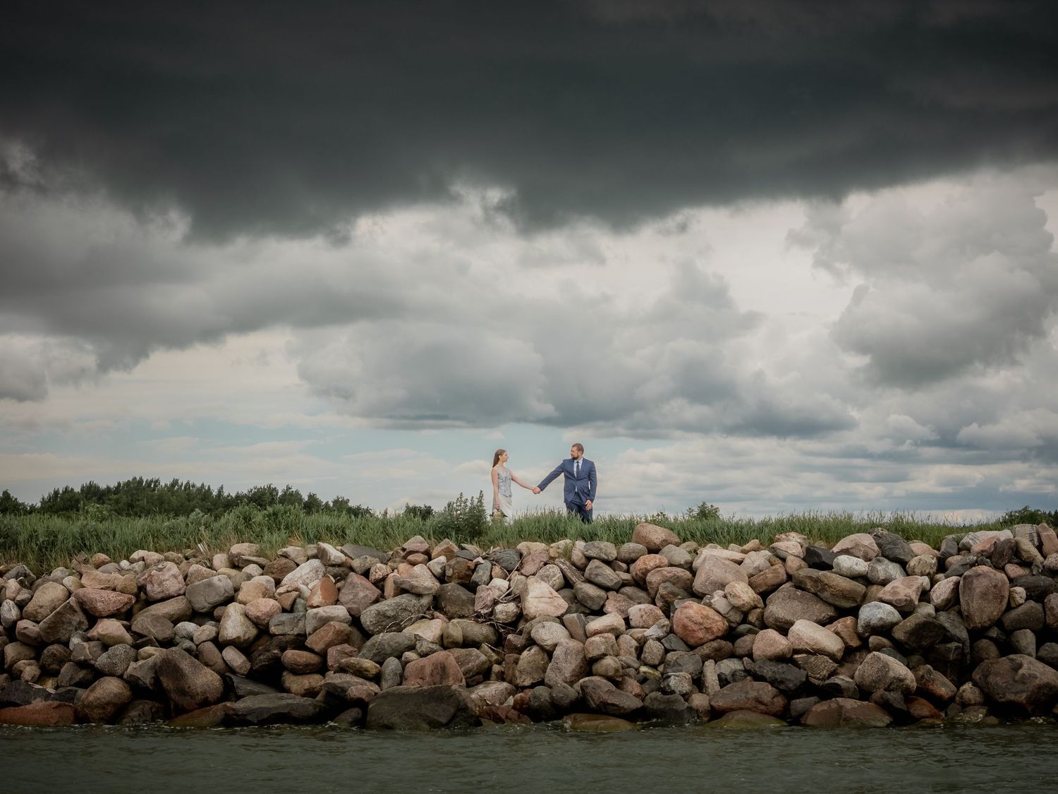 Couple in wedding attire walks on rocky shoreline under dramatic stormy sky with dark clouds and green landscape.
