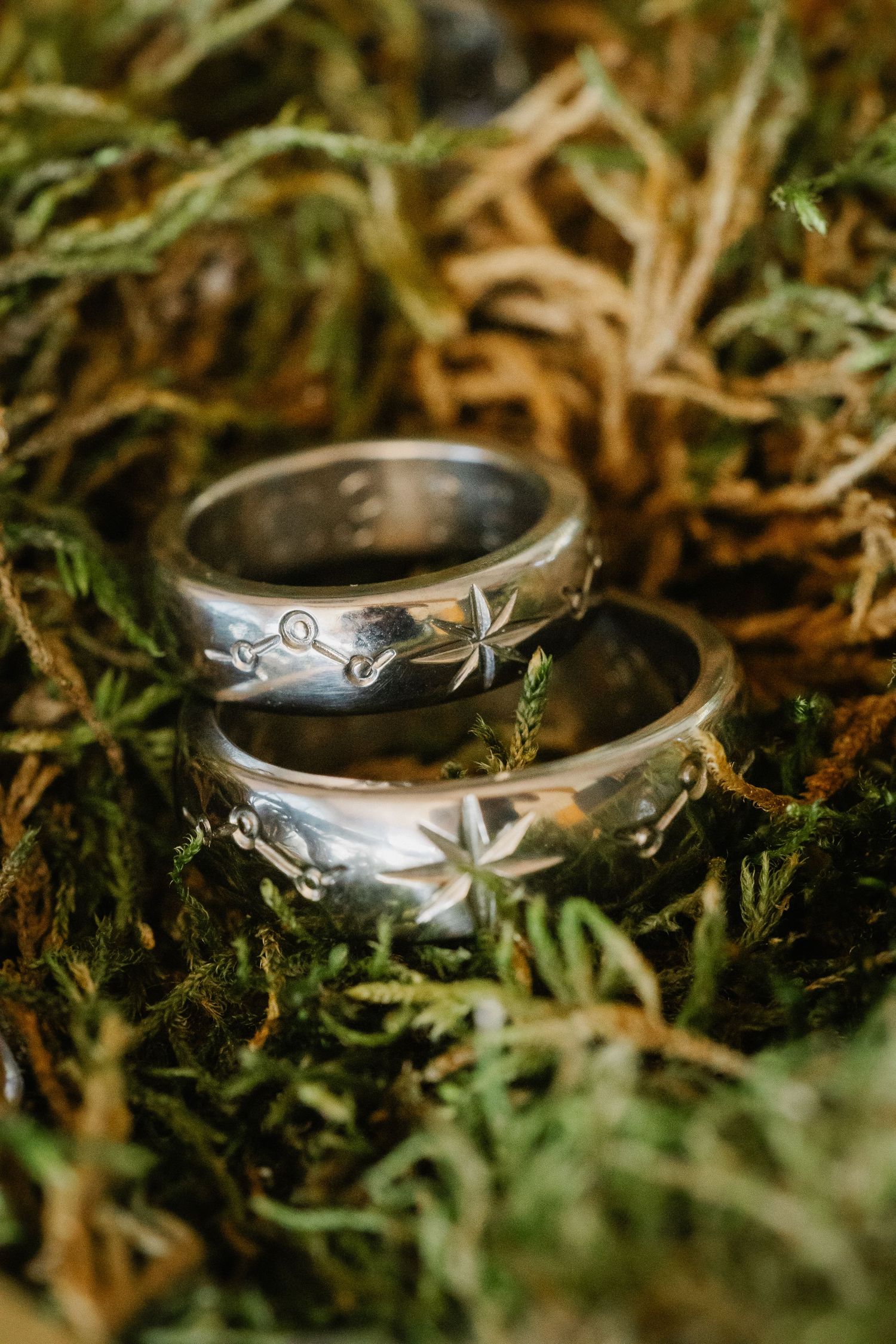 Two silver wedding bands with star engravings resting on green moss, perfect for nature-inspired weddings.