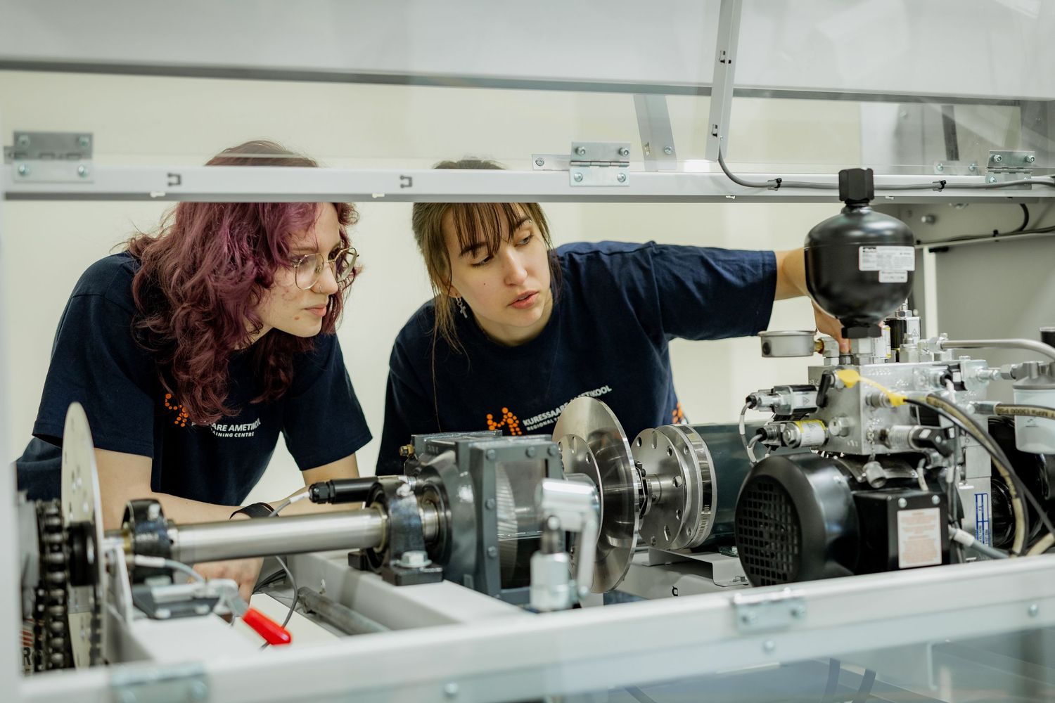 Two female engineering students in navy shirts inspect complex machinery components in a technical workshop.