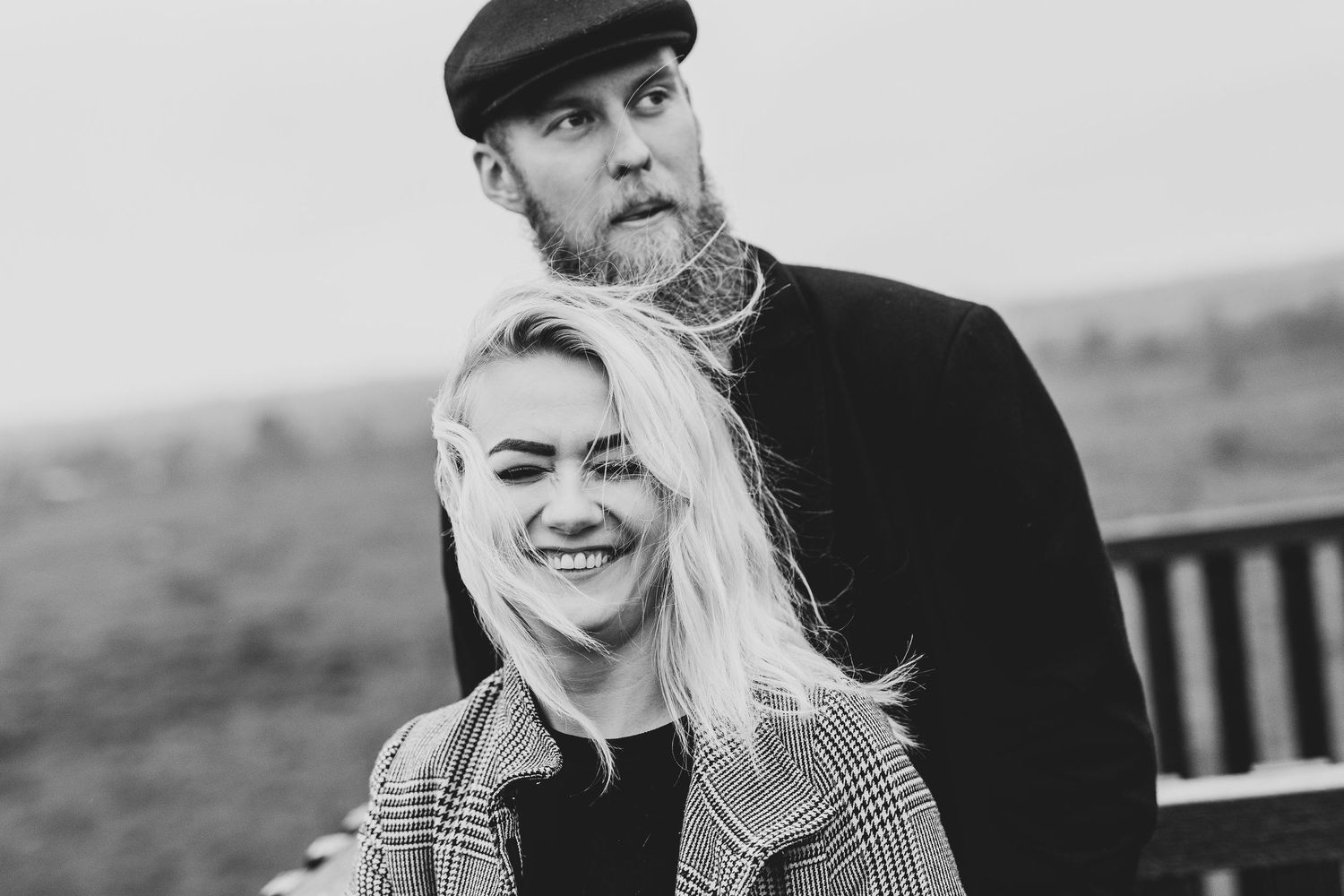 Black and white portrait of smiling blonde woman and tattooed bearded man outdoors on overcast day.