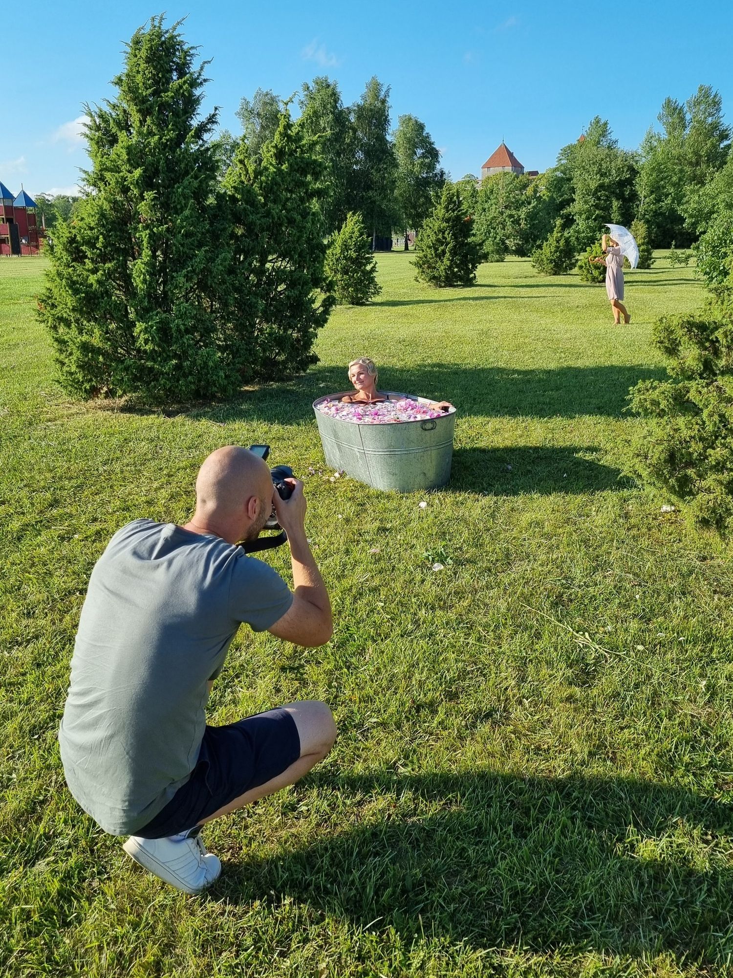 Bald photographer crouching on grass photographing a woman sitting in a flower-filled metal tub outdoors.