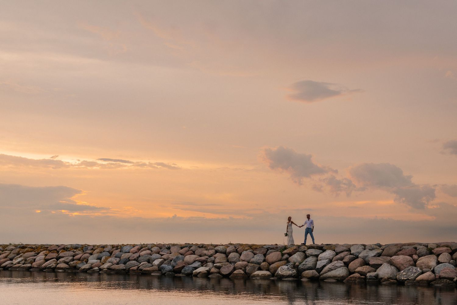 Couple holding hands on rocky jetty at golden sunset over calm water.