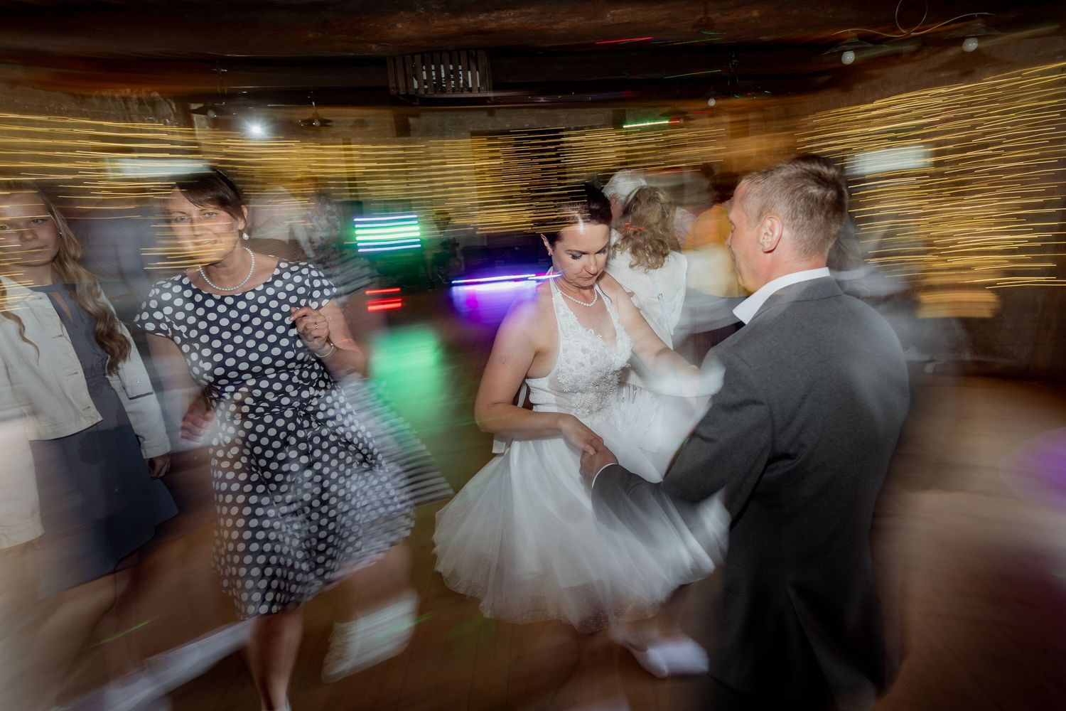 Bride and groom dancing with guests at a lively wedding reception, captured with motion blur effect.