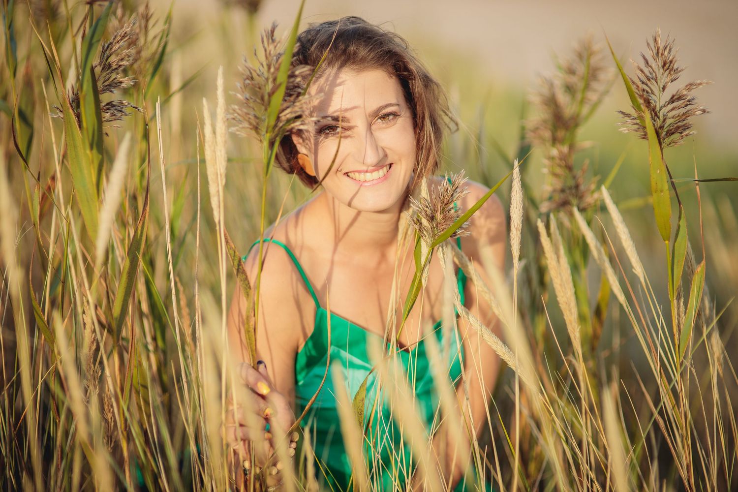 Smiling young woman in green dress surrounded by tall grass and wheat stalks in a sunny field.