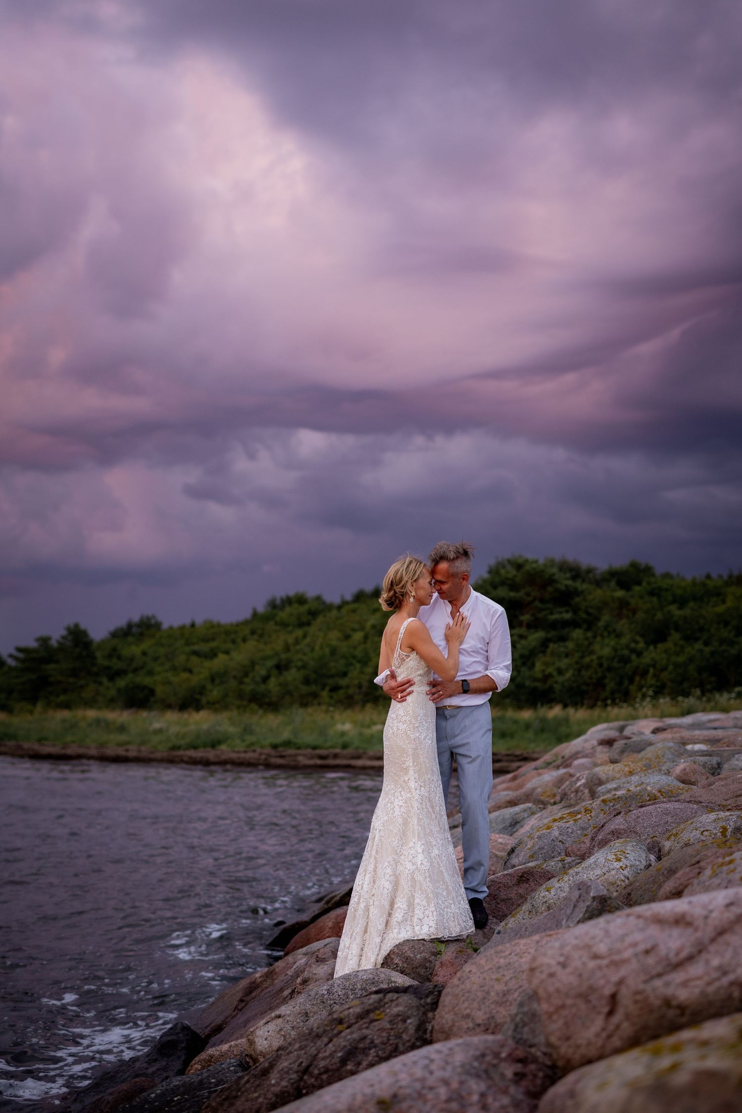 Bride and groom embrace on rocky shoreline under dramatic purple storm clouds at sunset.