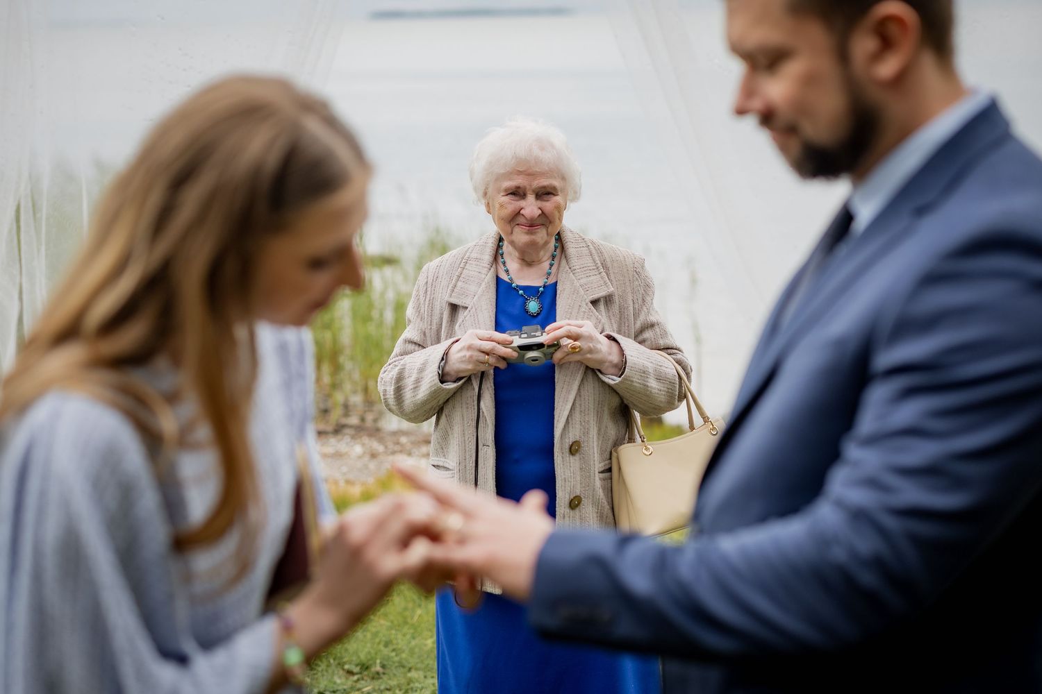 Elderly woman smiling and photographing a young couple exchanging rings at an outdoor wedding ceremony.