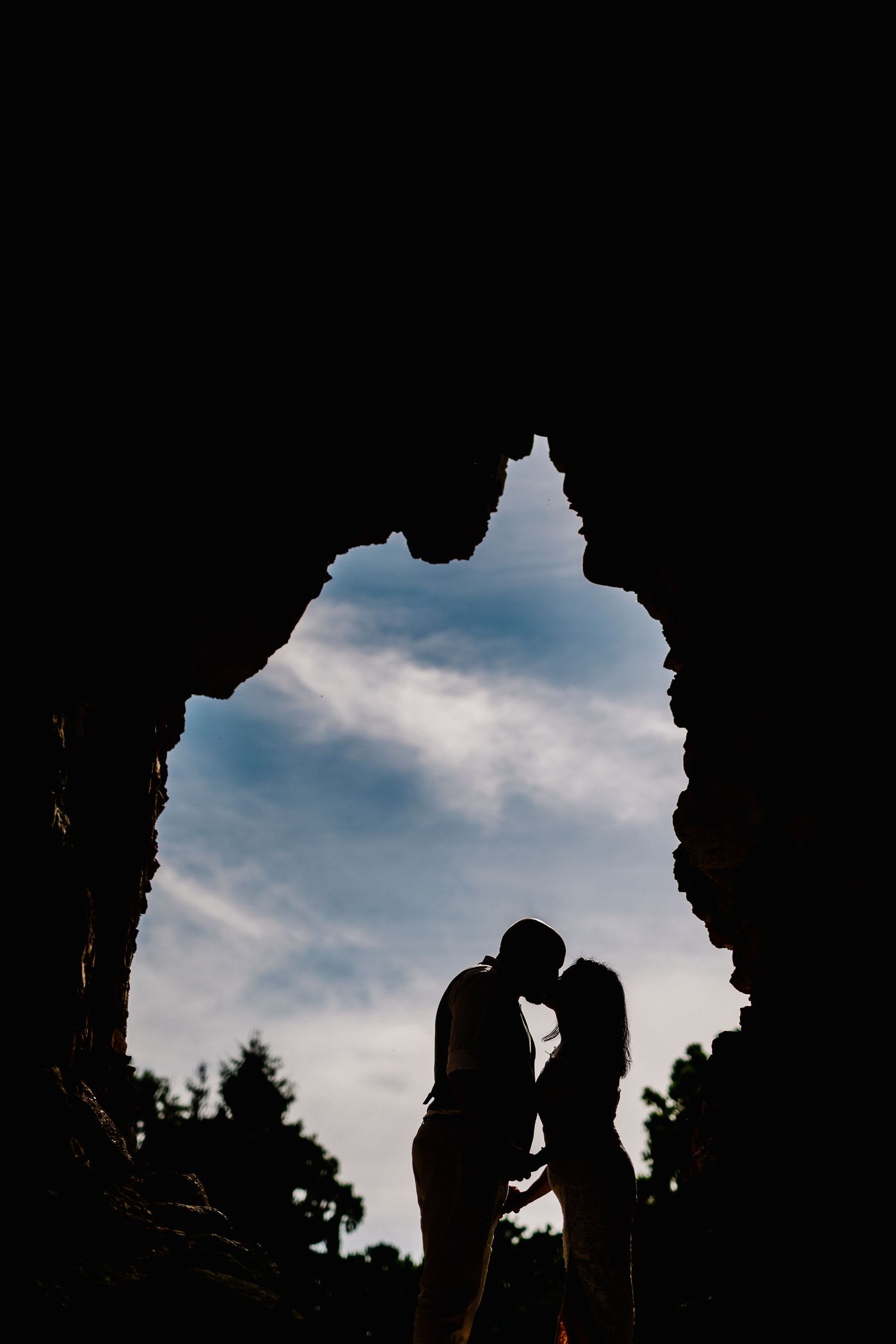 Silhouette of a couple kissing framed by a rocky cave opening with a blue sky backdrop.