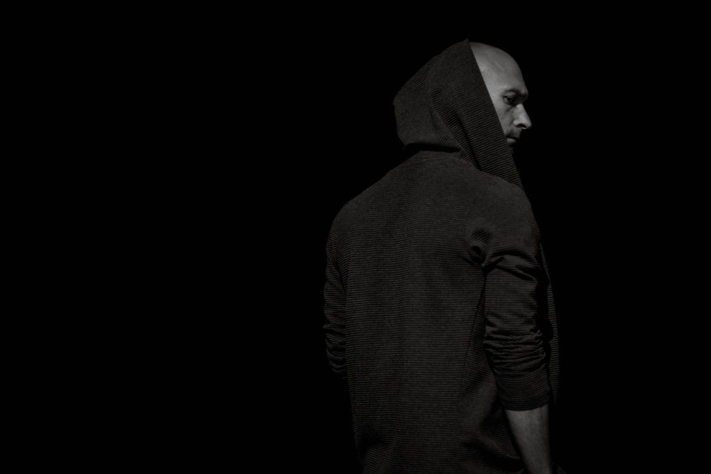 Photographer Valmar in a black hoodie stands against a dark background in a dramatic black and white portrait.