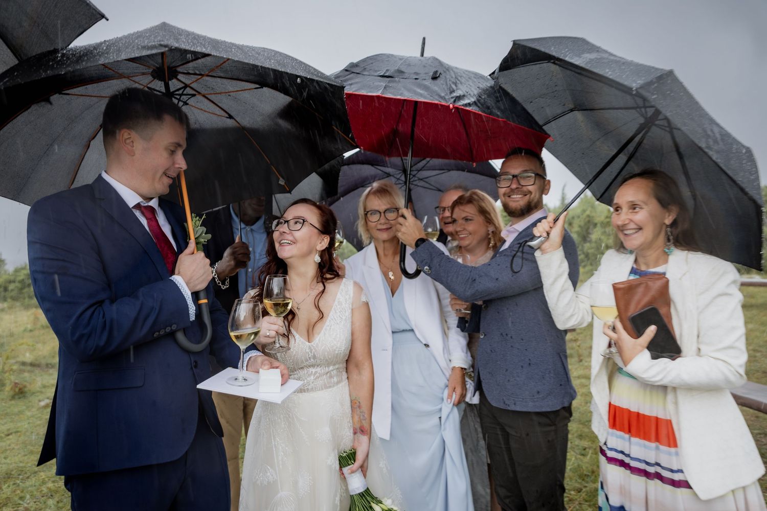 Wedding party sheltering under black umbrellas in the rain during an outdoor ceremony with bride in white gown.