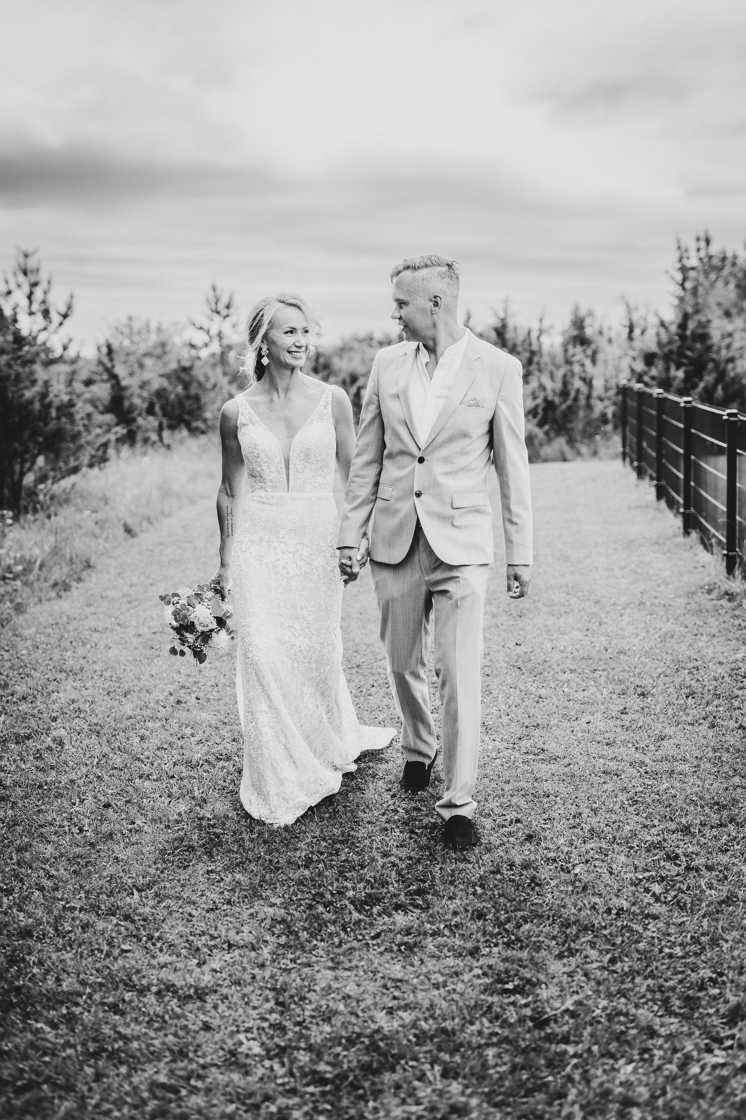 Black and white photo of a smiling bride and groom walking hand in hand on a grassy path on their wedding day.