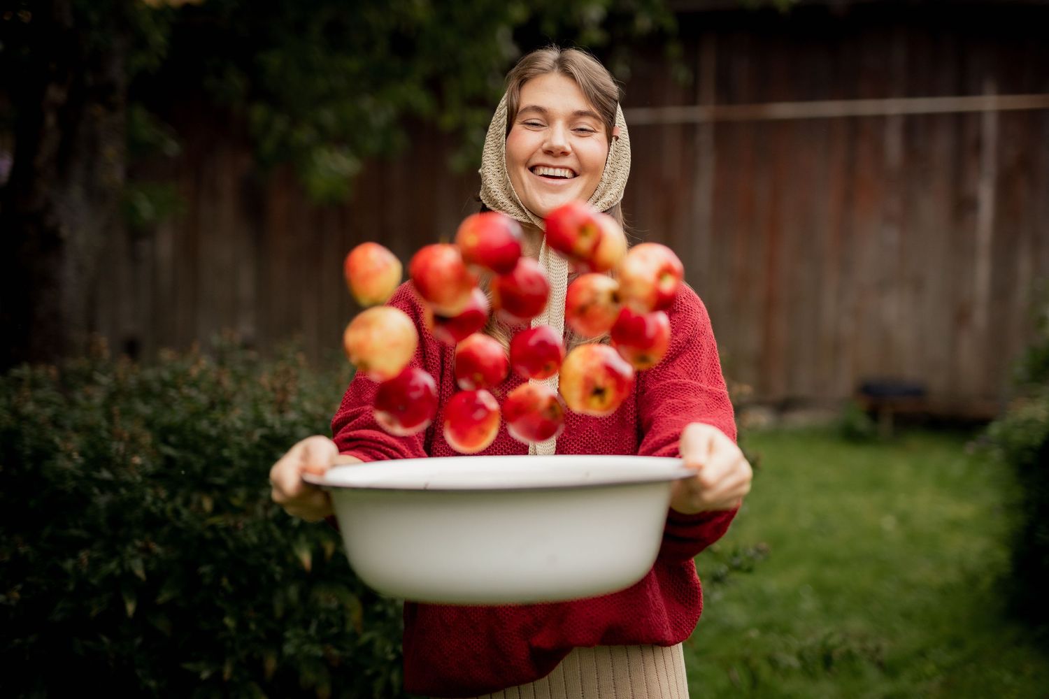 Smiling woman in red sweater and headscarf tossing fresh red apples in a white basin outdoors.