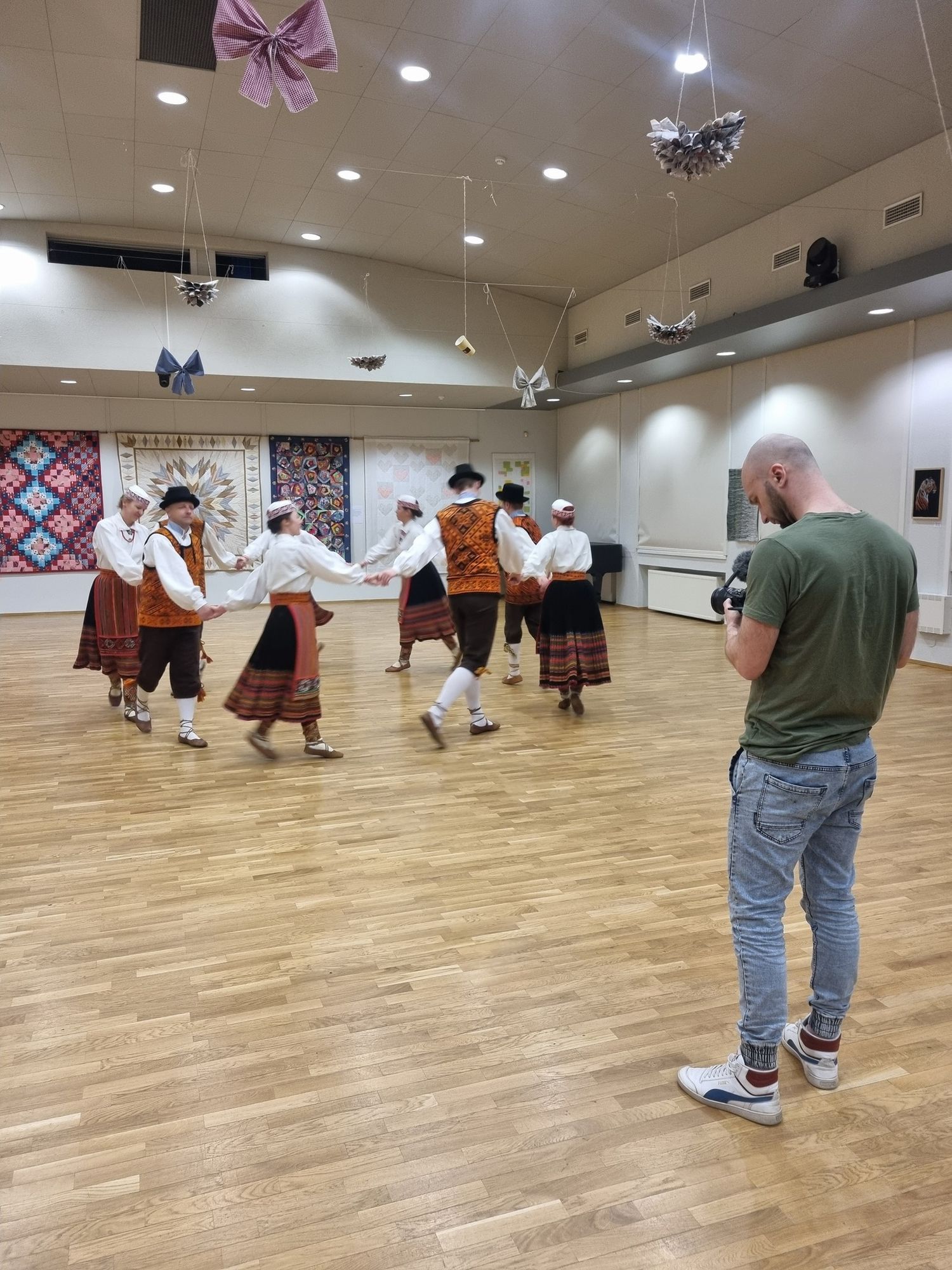 Photographer documenting traditional folk dancers in colorful costumes performing in a bright dance hall.