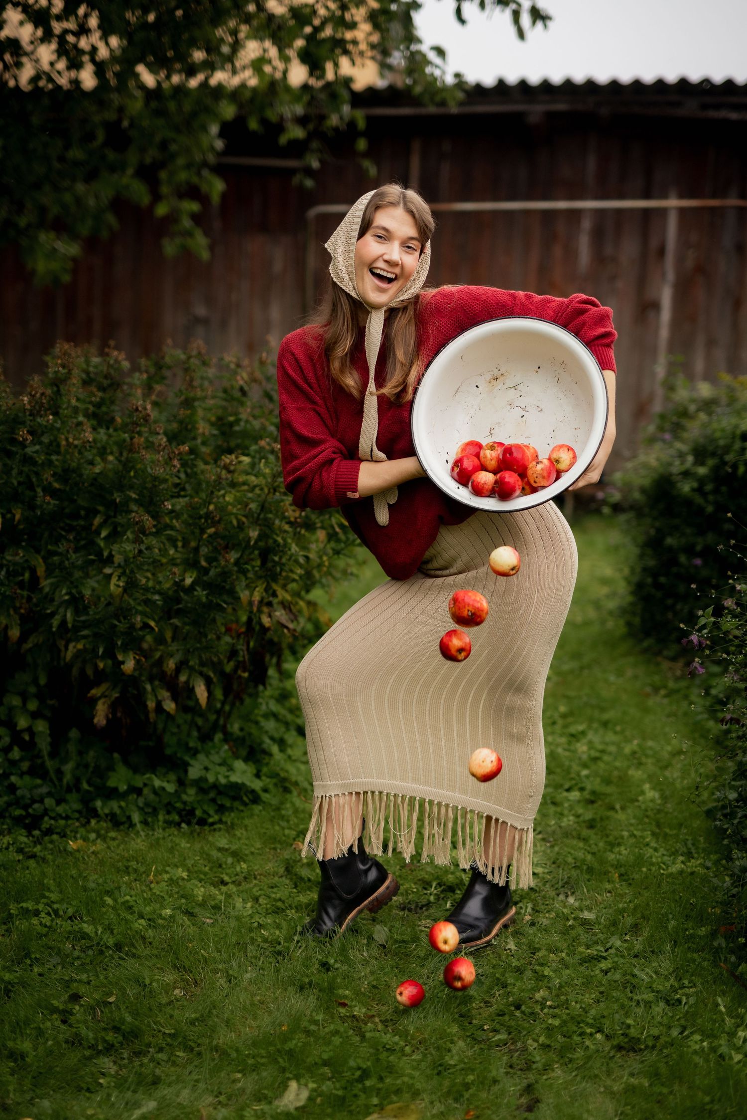 Woman in red jacket and headscarf laughing while dropping red apples from a large bowl in a garden.