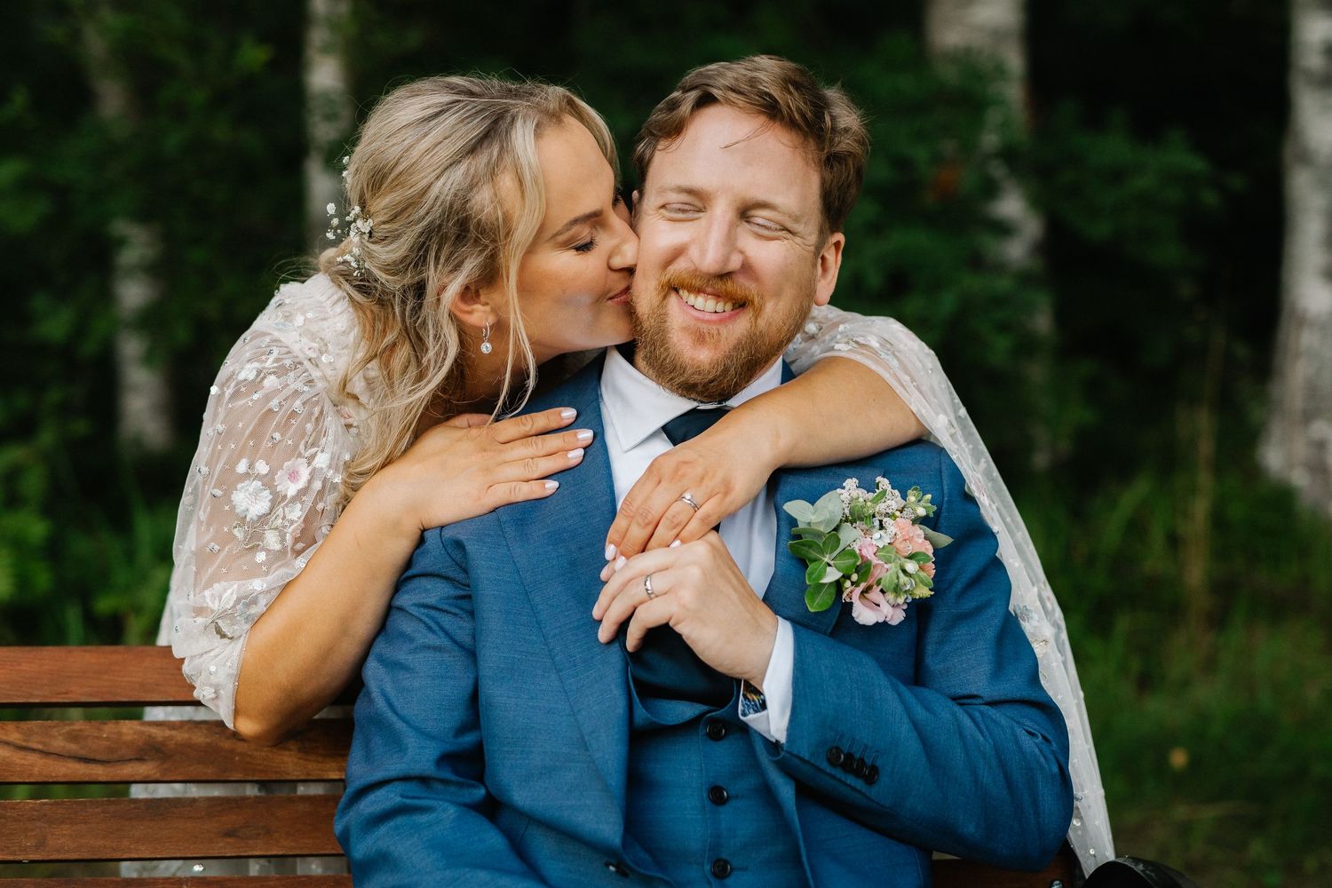 Bride kissing groom on cheek while he smiles, seated on bench, groom in blue suit with floral boutonniere.