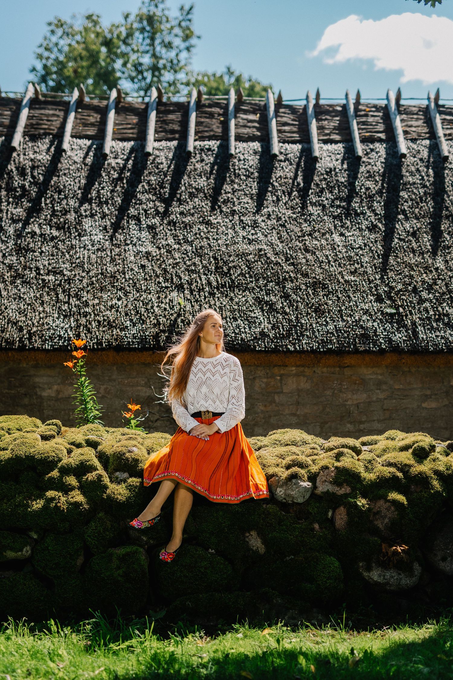 Woman in orange skirt and white blouse sitting on mossy stone wall beside thatched roof cottage on sunny day.