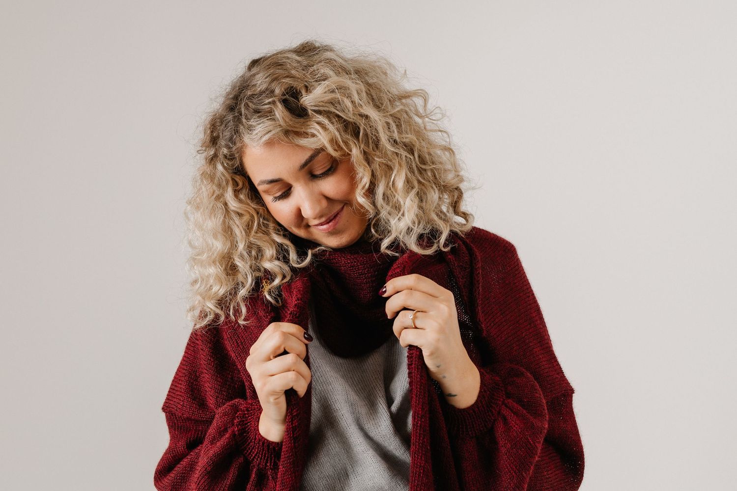 Smiling woman with curly blonde hair wearing a burgundy coat and gray sweater against a white background.