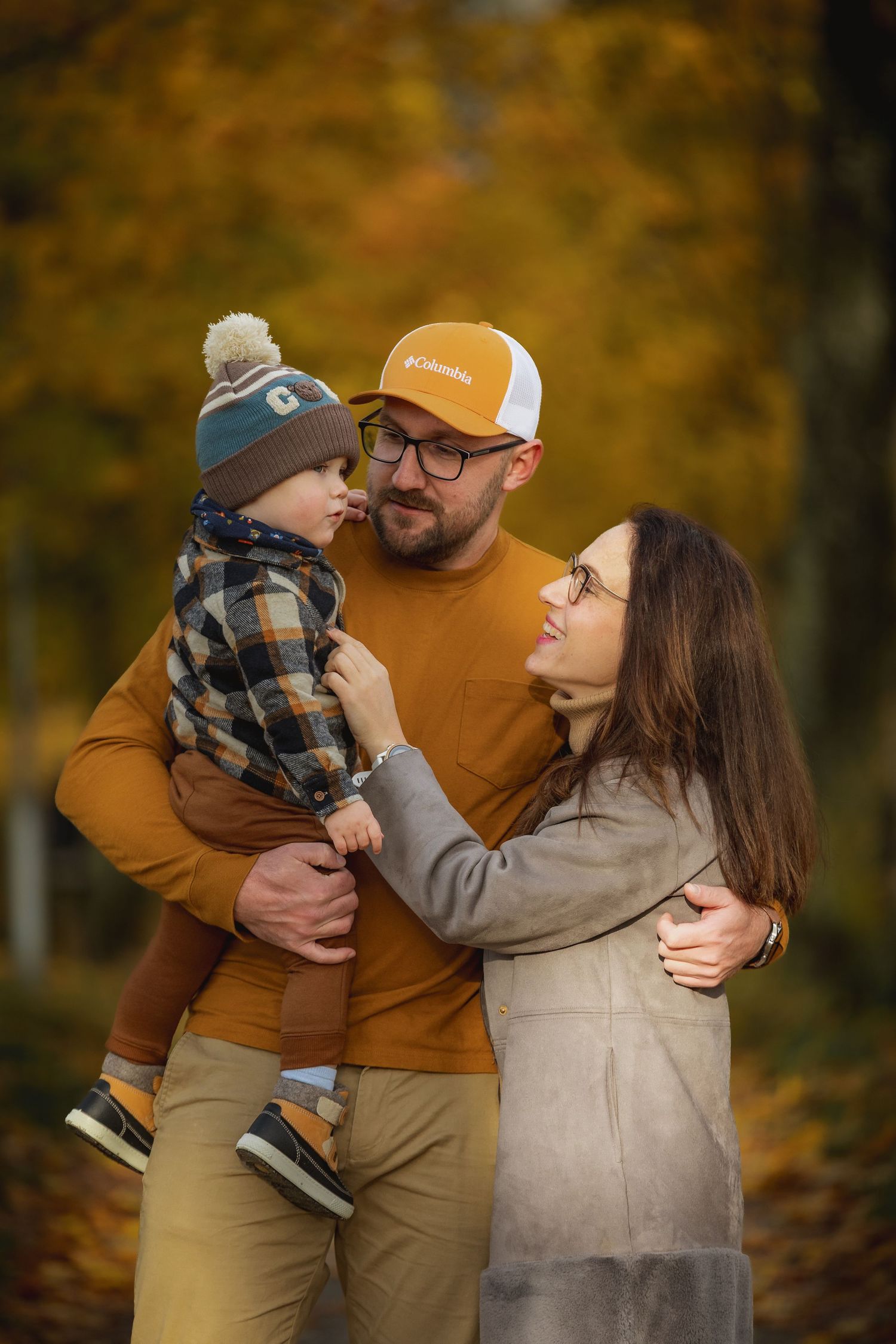 A family of three enjoys an autumn forest walk, with dad holding toddler in plaid jacket and mom in gray coat smiling warmly.