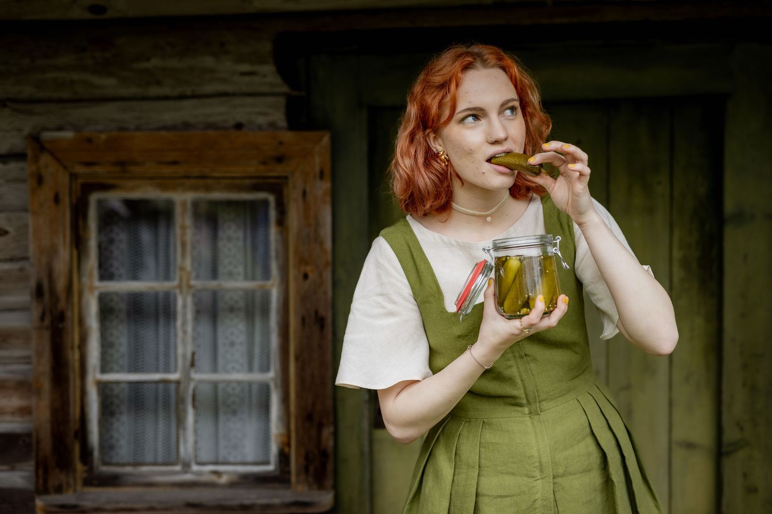 Red-haired woman in green apron eating a pickle from a jar outside a rustic wooden cabin.