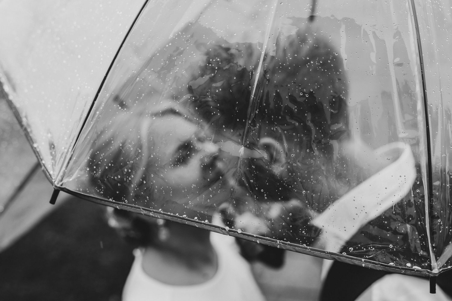 Black and white photo of a couple kissing under a clear umbrella in the rain on their wedding day.