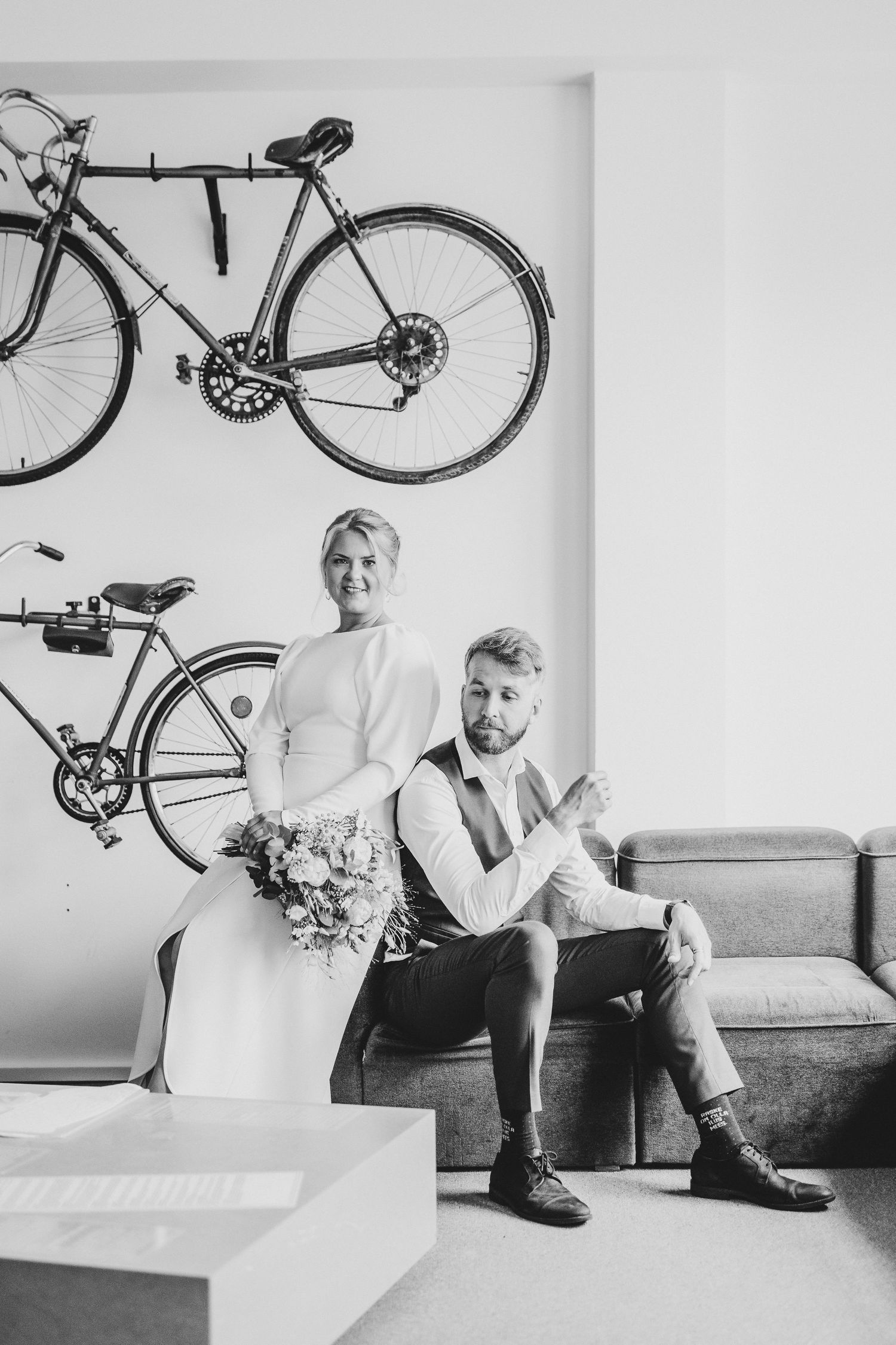 Black and white photo of a bride holding flowers standing behind a groom seated on a sofa with bicycles mounted on the wall.