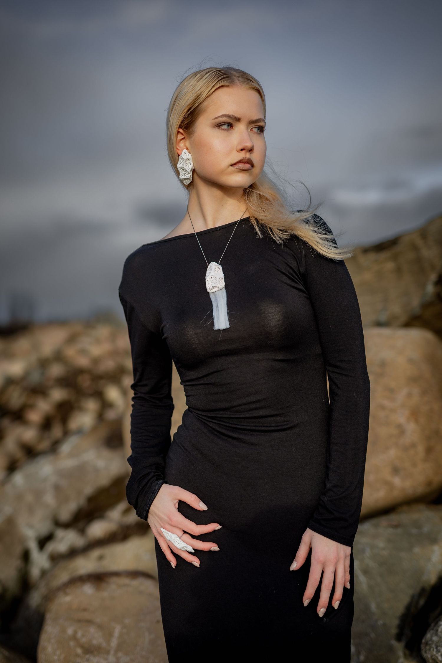 Blonde woman in elegant black long-sleeve dress poses on rocky terrain wearing statement white jewelry.