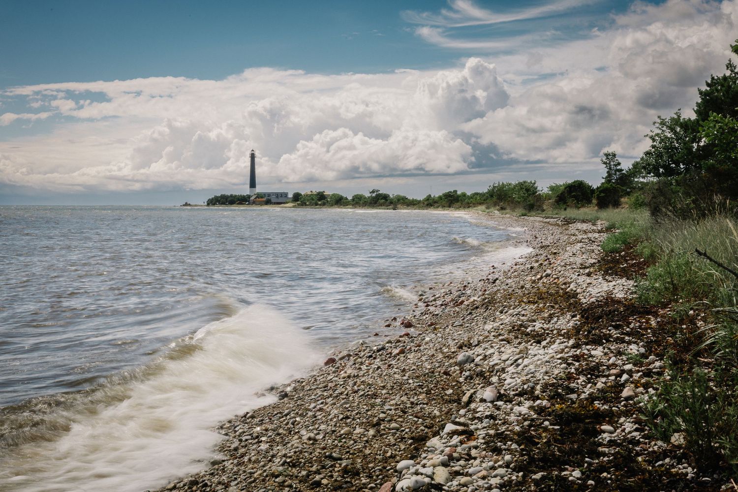 Rocky shoreline with gentle waves leading to a distant lighthouse under dramatic cloudy skies on a serene coastal landscape.