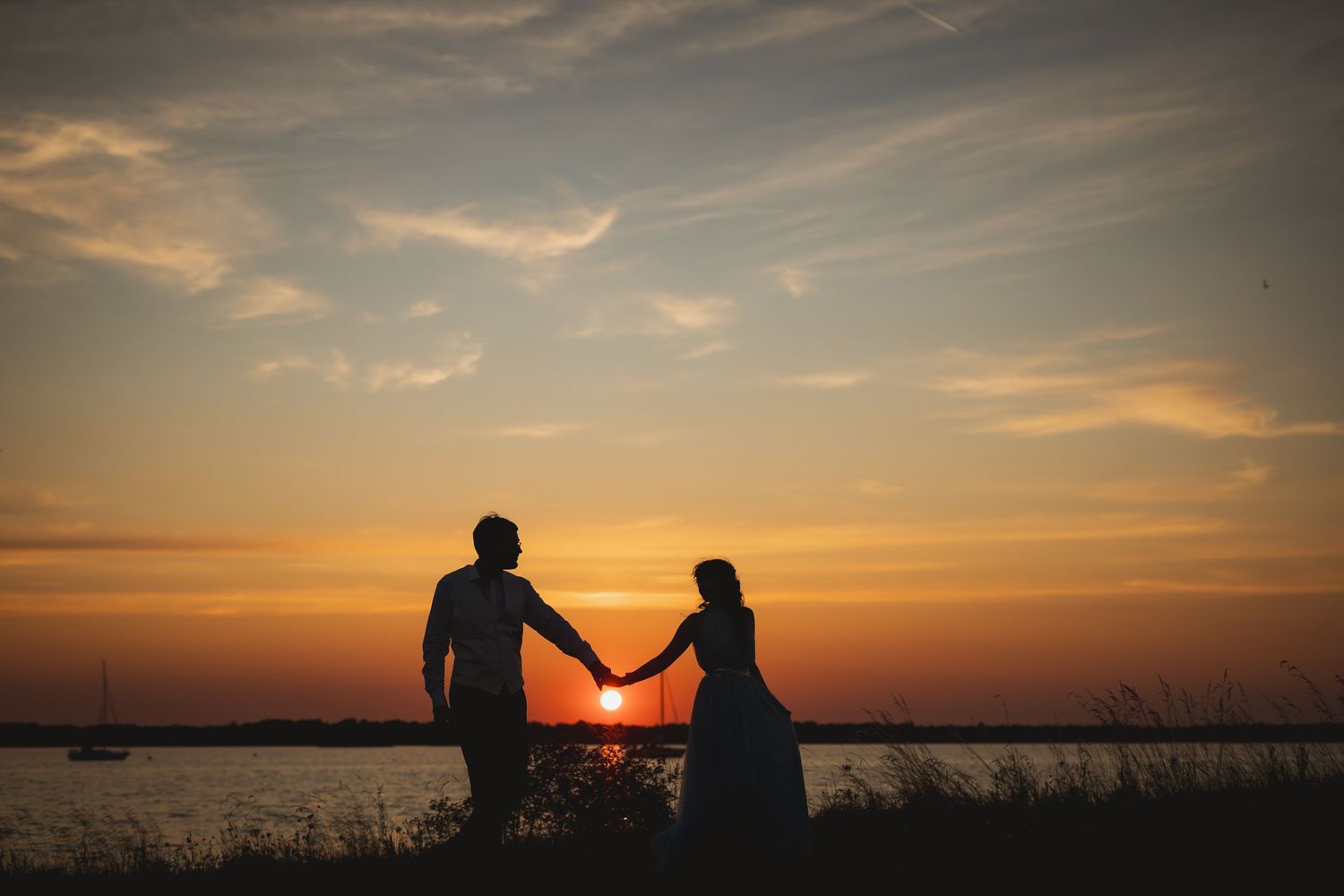 Silhouette of a couple holding hands at sunset by the water, with a vibrant orange sky in the background.