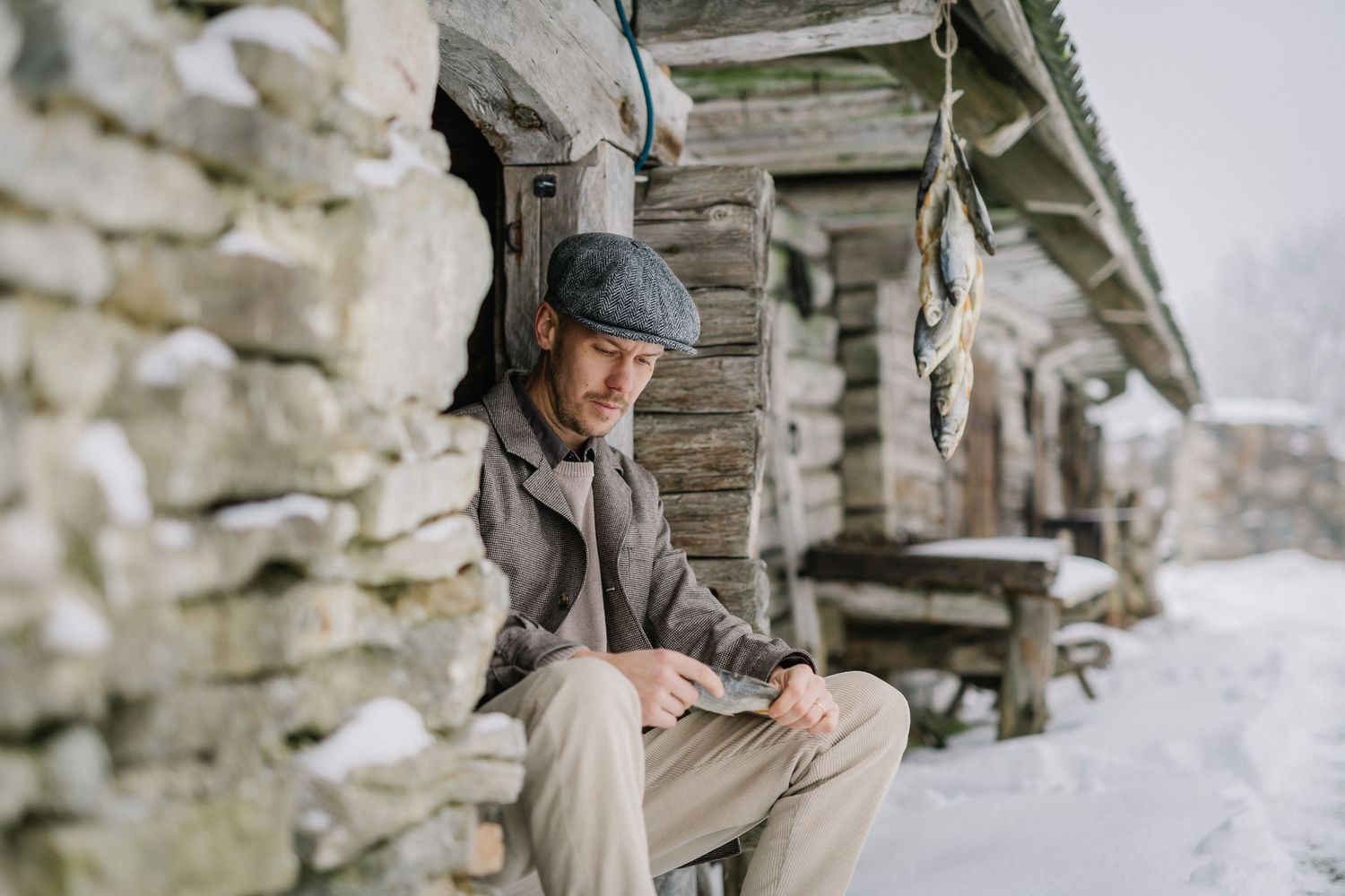 Man in flat cap reads outdoors by rustic log cabin in snowy winter landscape.