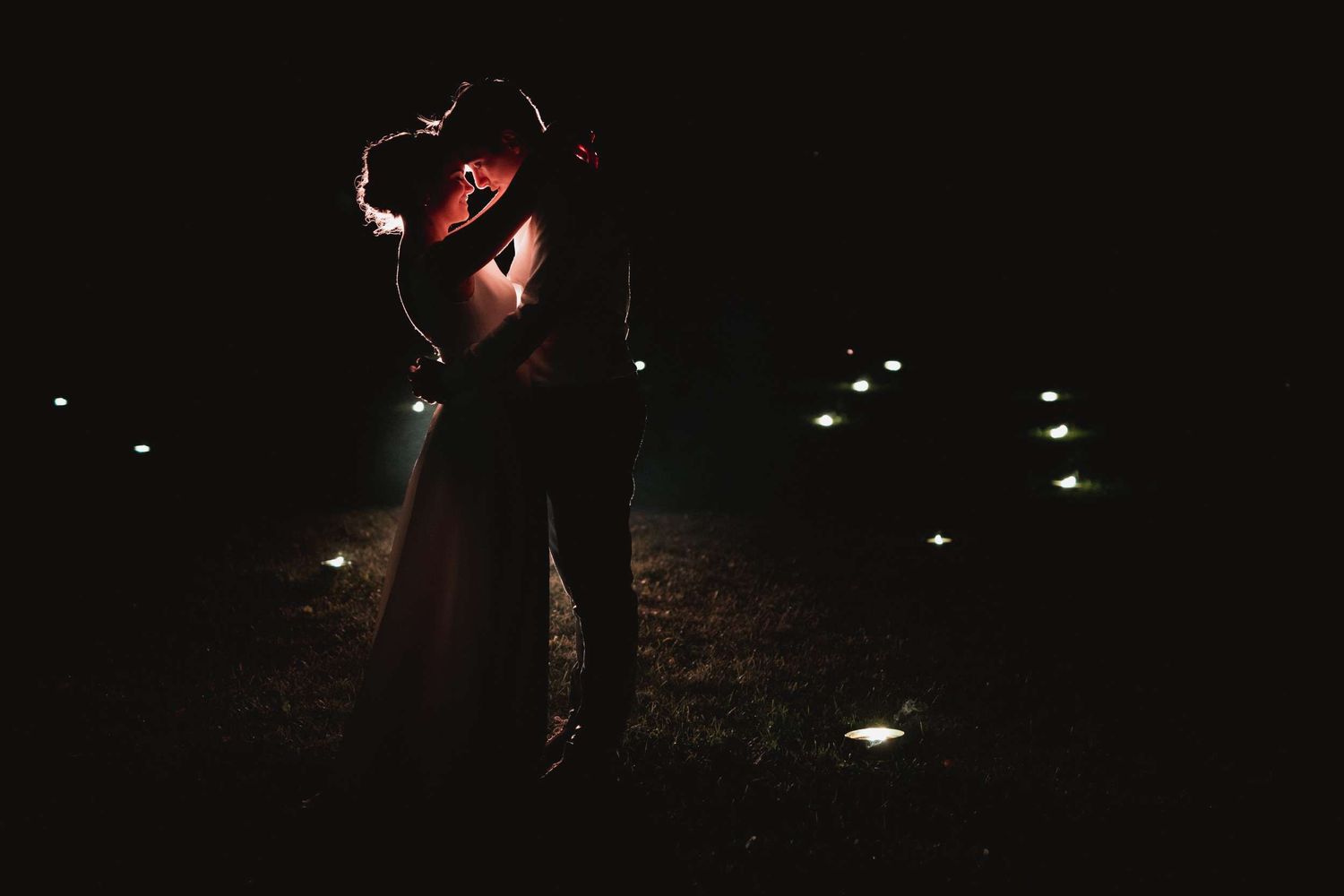 A romantic couple embraces in a tender nighttime silhouette illuminated by soft ground lights.