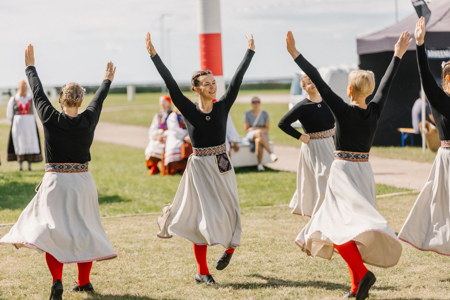 Traditional folk dancers in black tops and white skirts perform an energetic outdoor cultural dance routine at a sunny festival event.